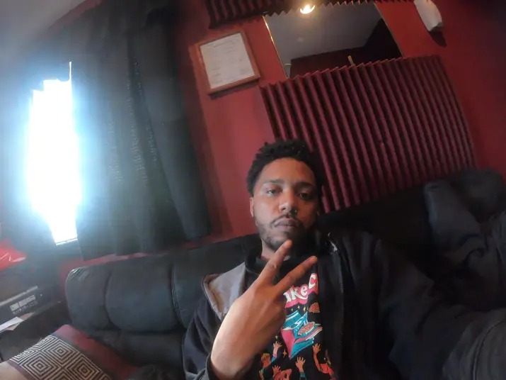 Young man with short curly hair making a peace sign, sitting on a black couch in a room with red walls and a window.