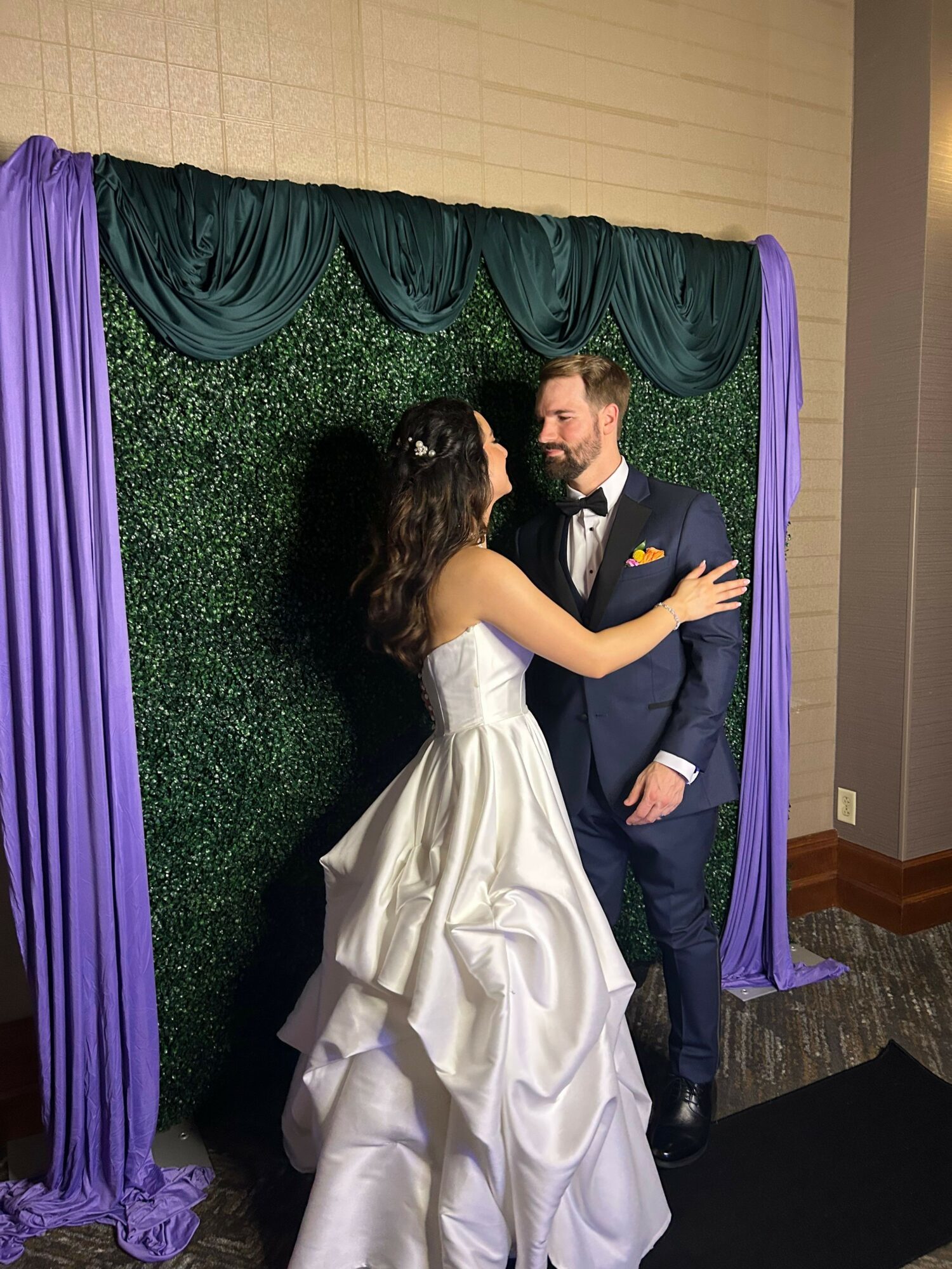 Two women are engaged in conversation in front of a green backdrop with purple curtains, one in a white dress and the other in a dark suit.