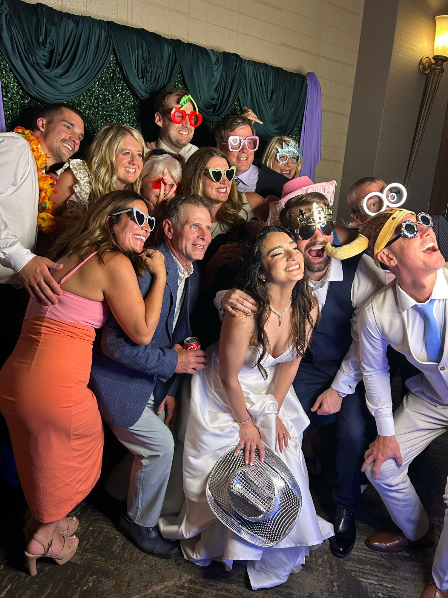 Group of people in costumes and formal attire, smiling and posing together indoors, some wearing masks and accessories.