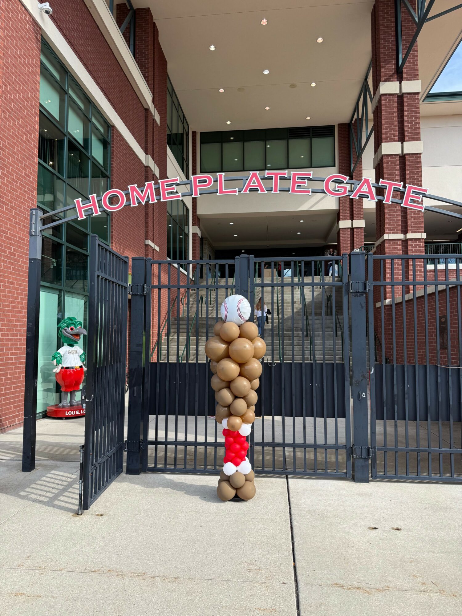 Giant cluster of brown and red balls on a stand outside a building, with a sign reading 'HOMEPLATEGATE' and a person in the background.