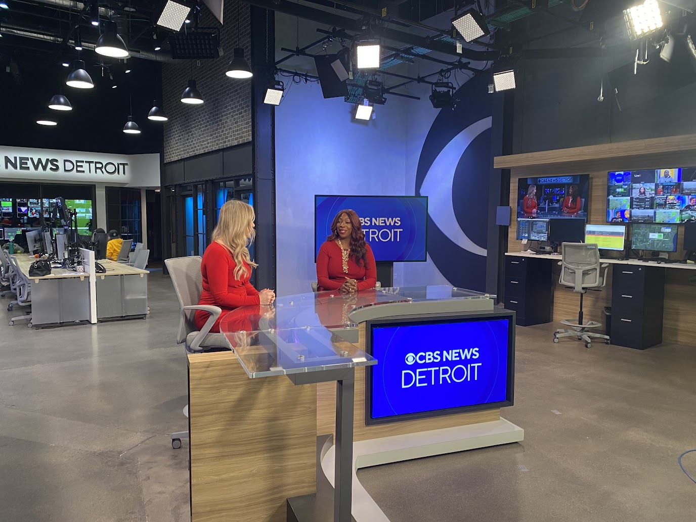 Two women in red outfits sit behind a news desk in a television studio with multiple monitors and a newsroom background.