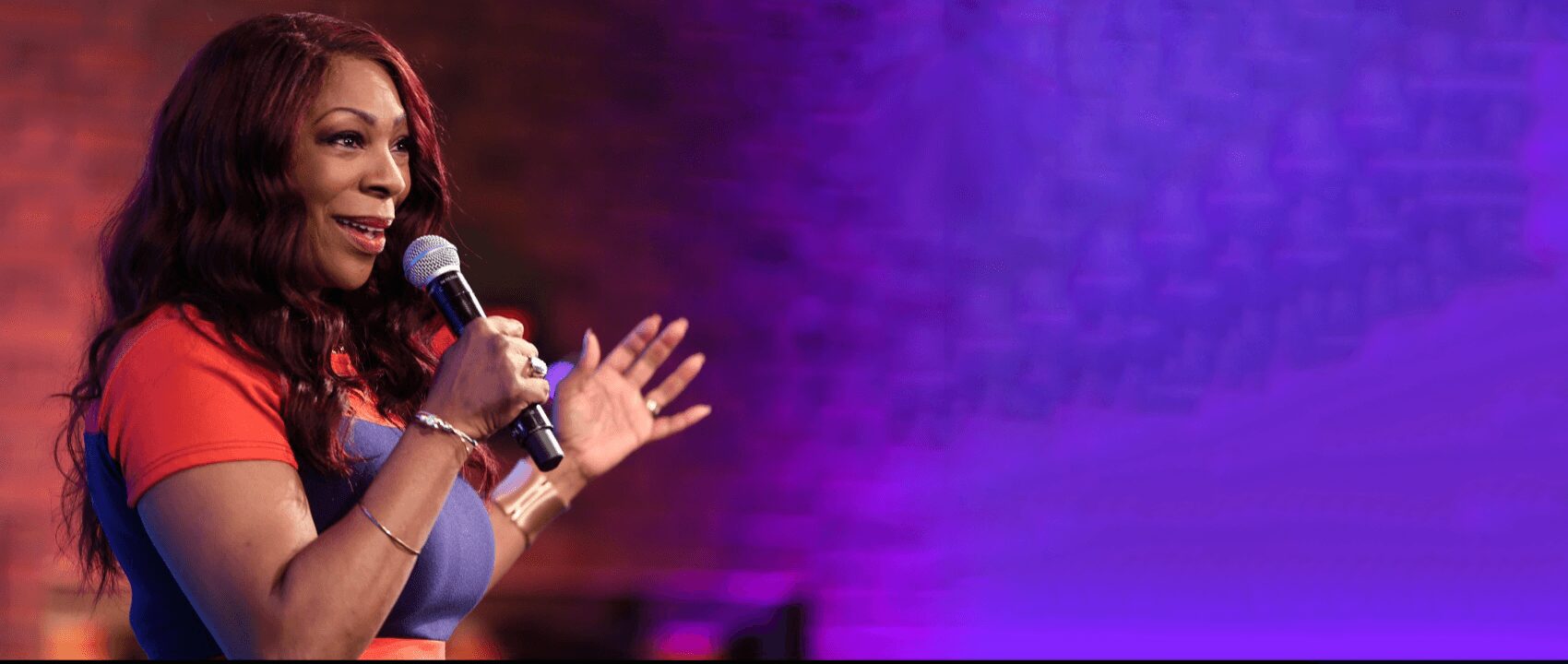 Woman speaking into microphone on stage with purple background, wearing red shirt, smiling, gesturing with hand.