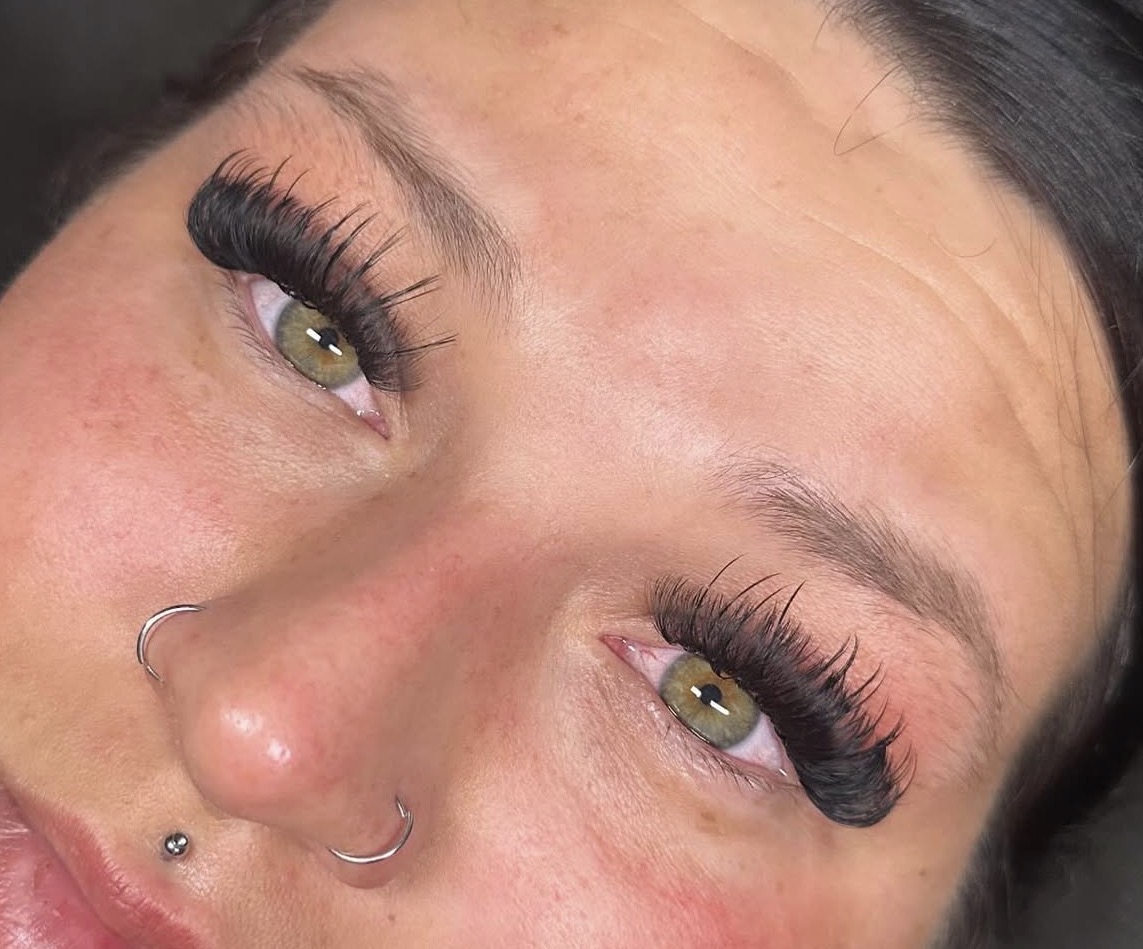 Close-up of a person's eyes with long eyelashes, light skin, and a nose piercing, looking upward.