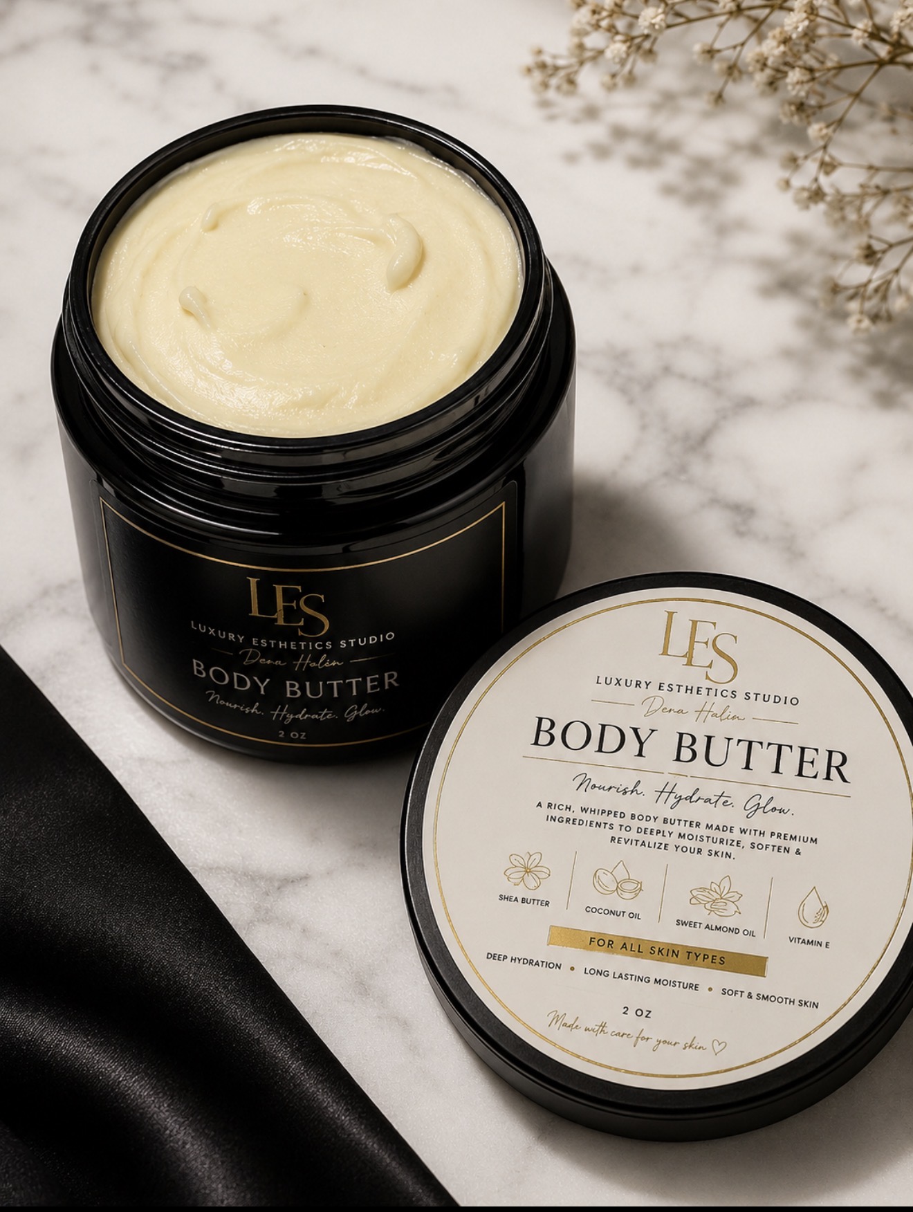 Open container of body butter on marble surface with lid beside, black fabric in foreground, dried flowers in background.