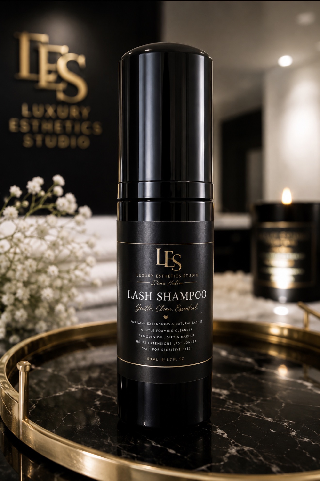 Black bottle of lash shampoo with gold and white text on a reflective surface, background includes blurred decor and flowers.