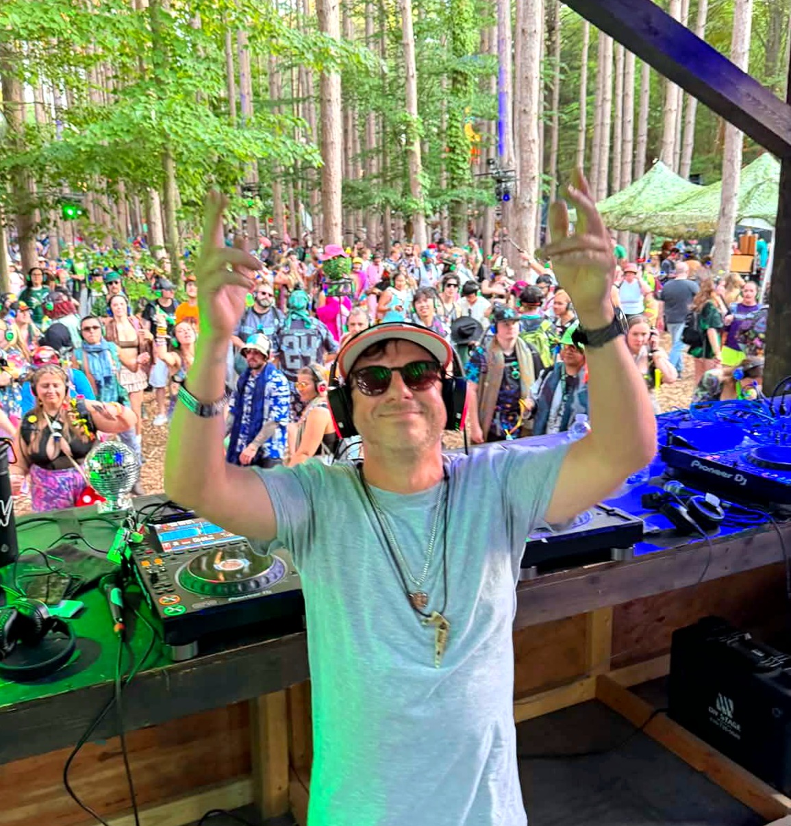 Person wearing sunglasses and headphones raising hands at outdoor event in forest, crowd in background.