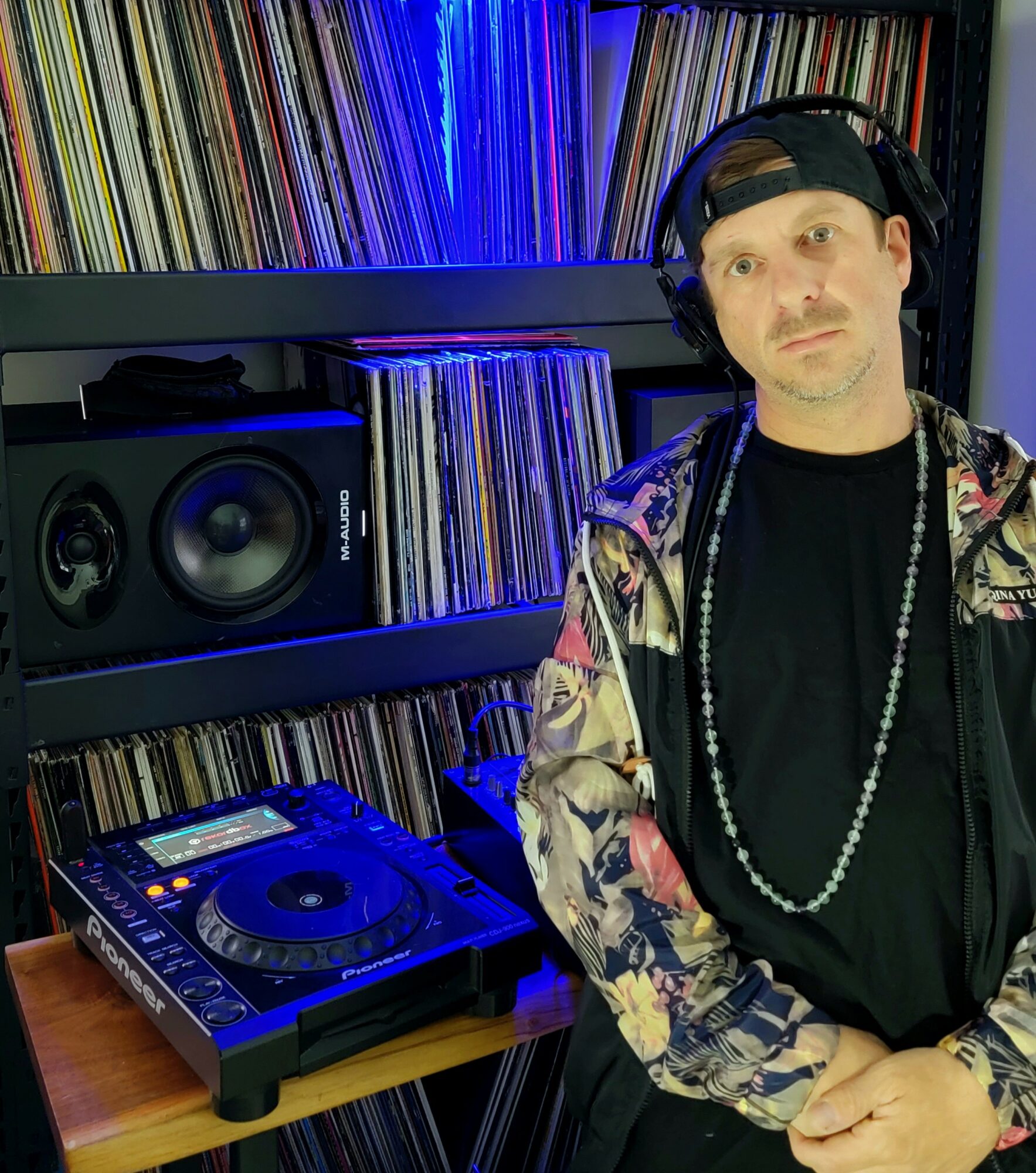 Person with headphones and a cap standing in front of shelves filled with vinyl records and a DJ turntable, blue lighting.