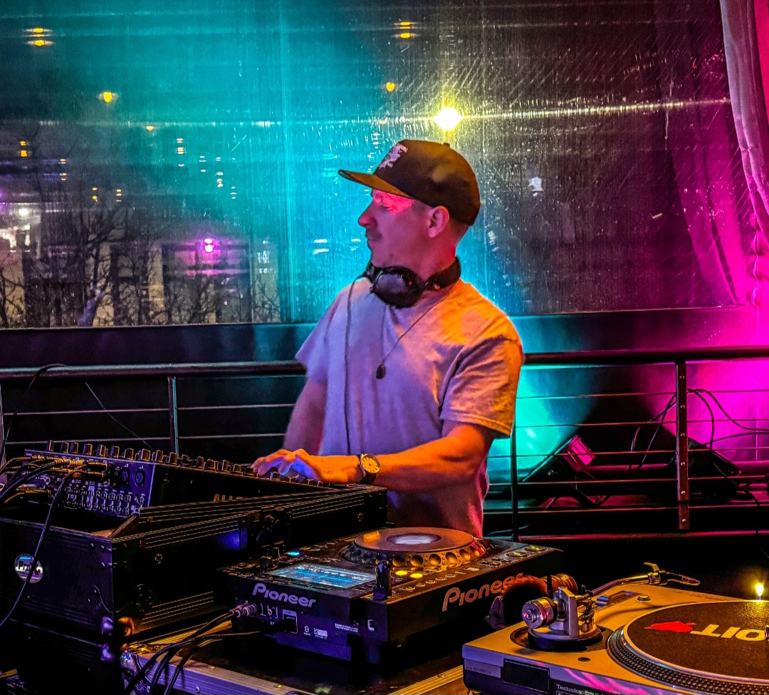 DJ wearing a cap and headphones, standing behind a mixing console with colorful lights in the background.