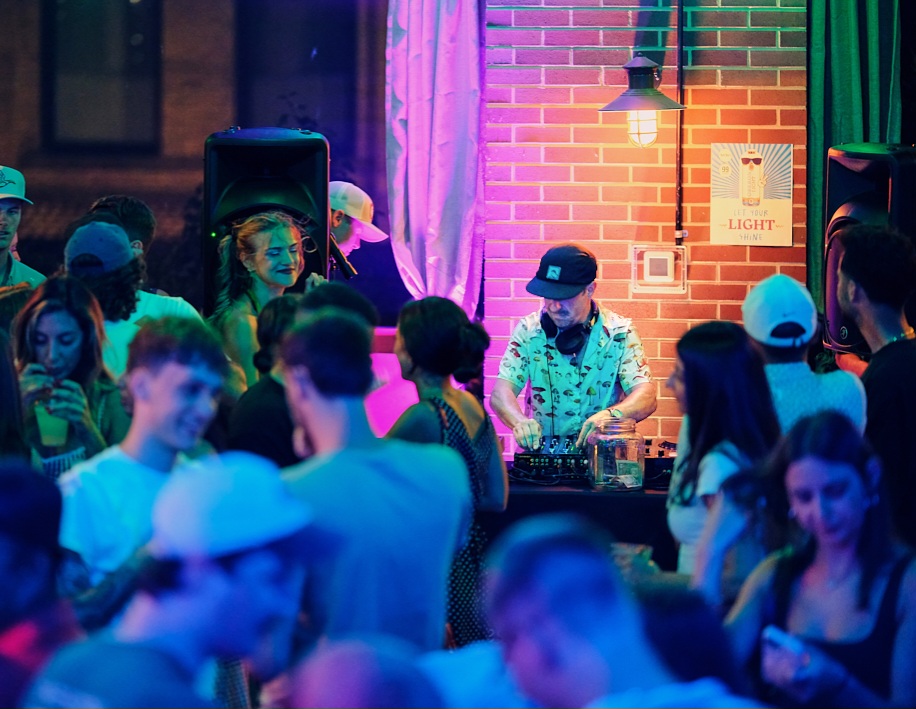 Crowd of people at a party with a DJ, colorful lighting, brick wall, and a sign that says 'Light'.
