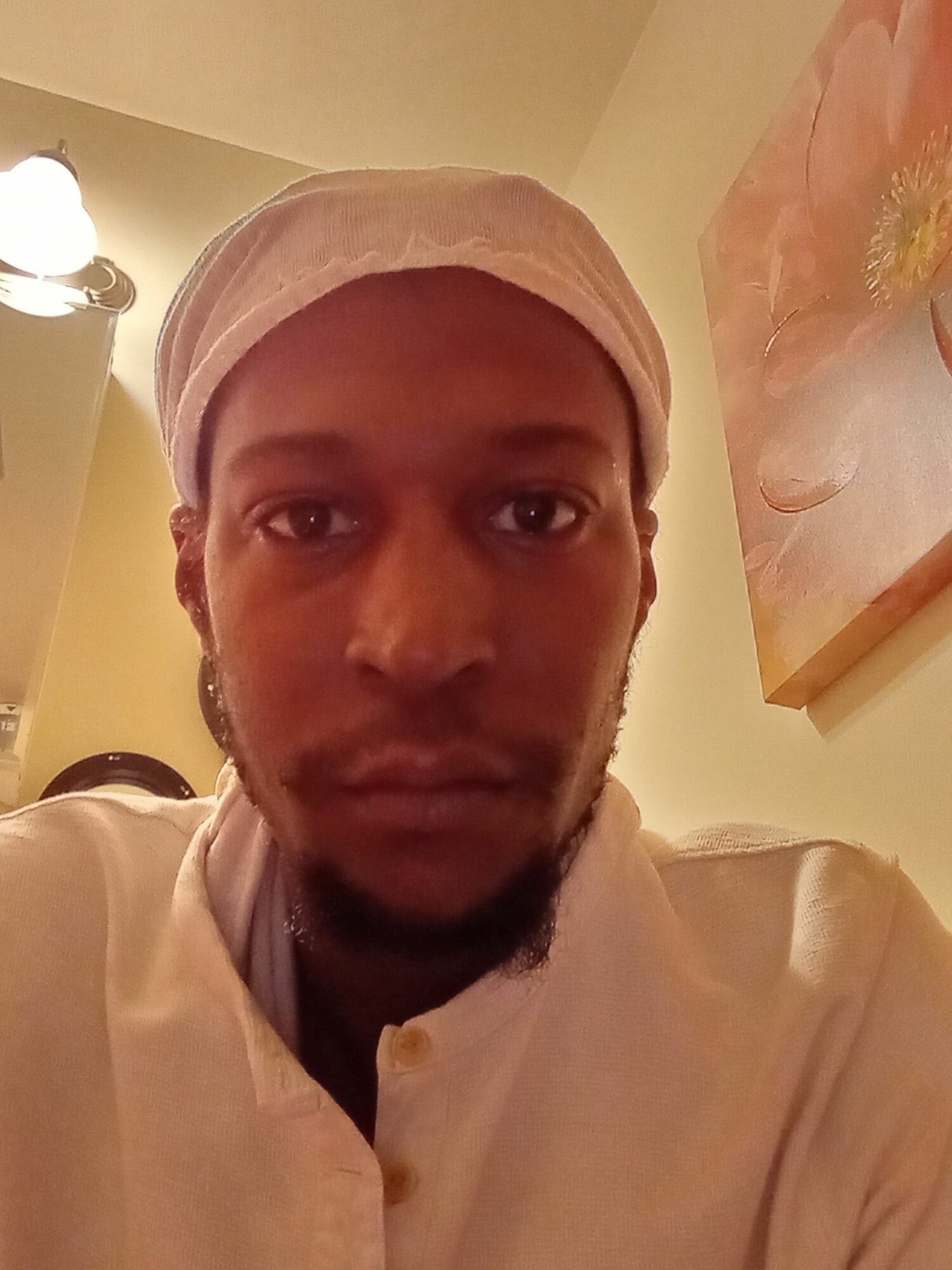 Person wearing a head covering, looking at the camera, with a light-colored background and a painting on the wall.