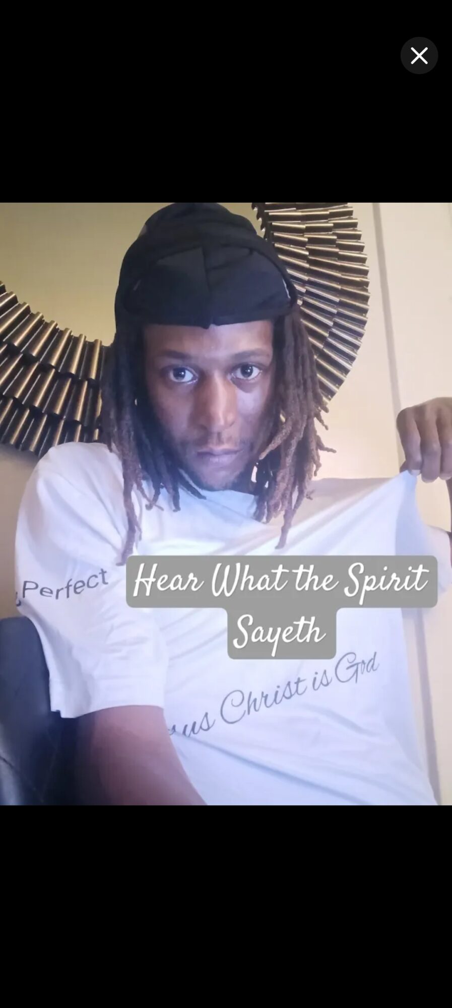 Person with dreadlocks wearing a black head covering, sitting indoors with a decorative circular wall piece behind them.