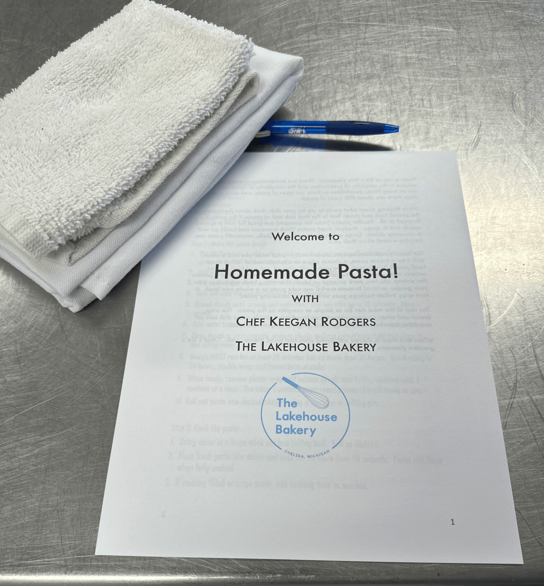 A printed menu titled 'Homemade Pasta!' with a logo for The Lakehouse Bakery, a pen, and a towel on a metallic surface.