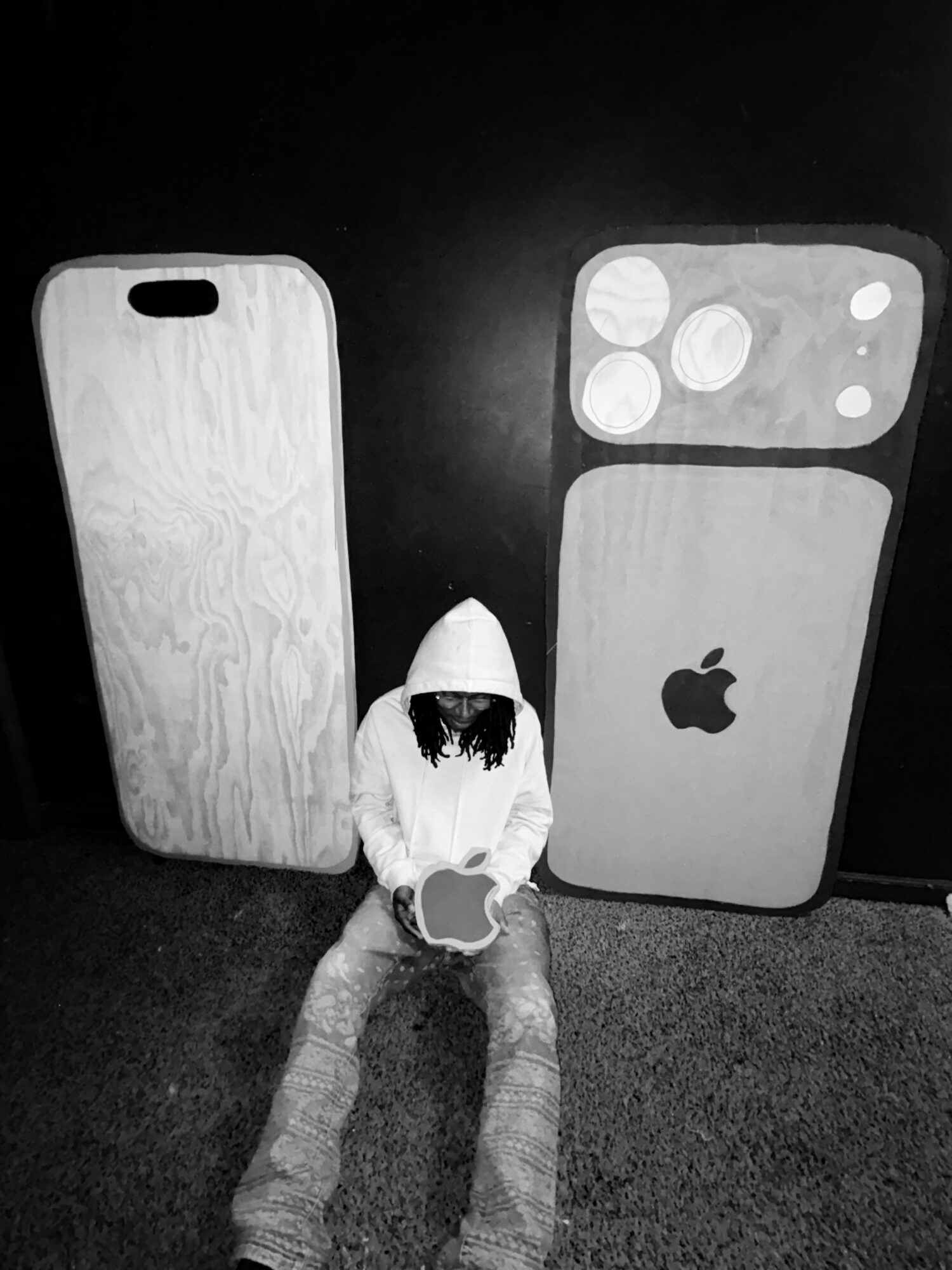 Person sitting on the floor with large iPhone and iPad images behind, person wearing hoodie and sunglasses, black background.
