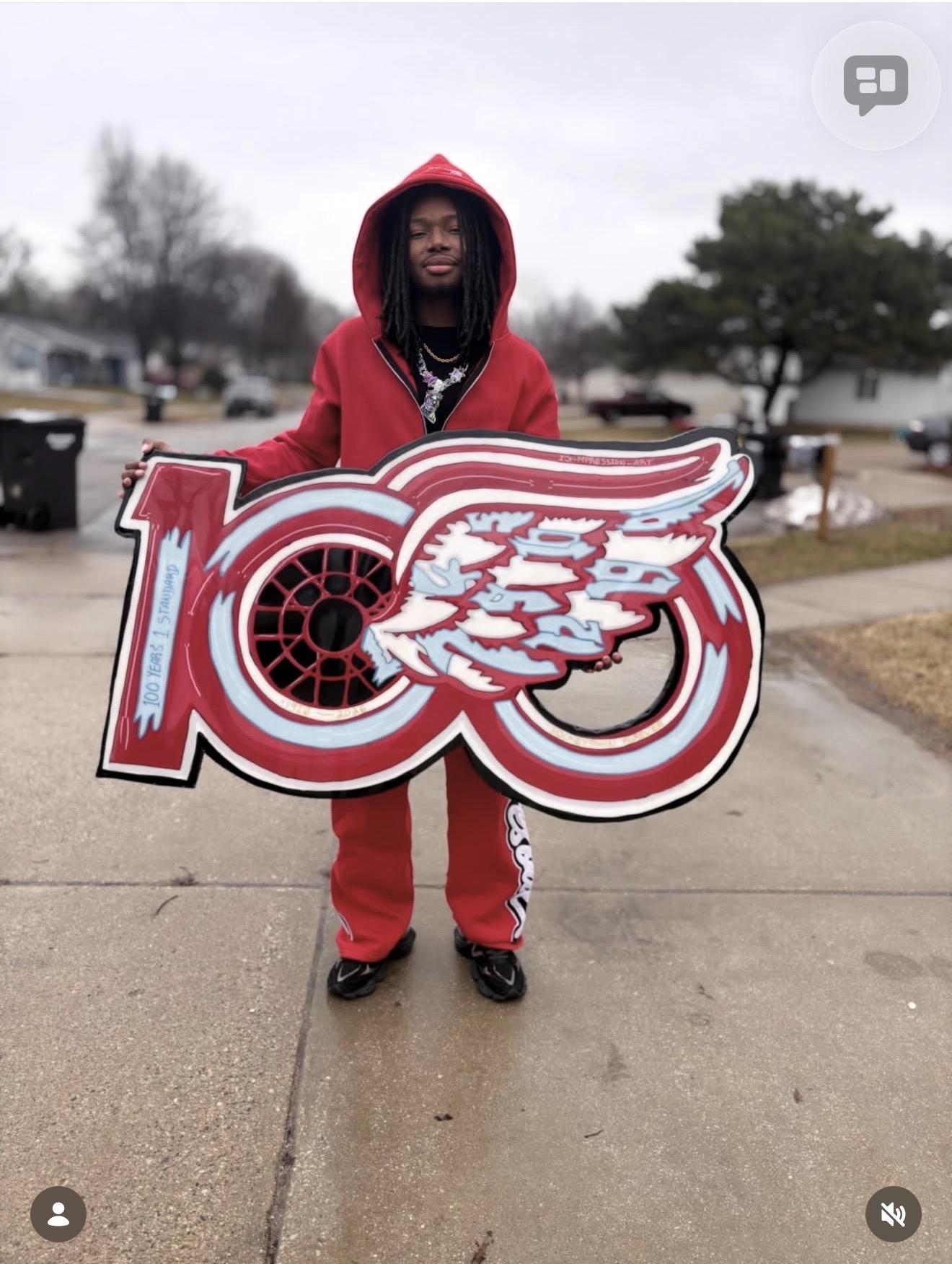Person in red hoodie and pants holding a large red and white sign with a winged logo, standing outdoors on a sidewalk.