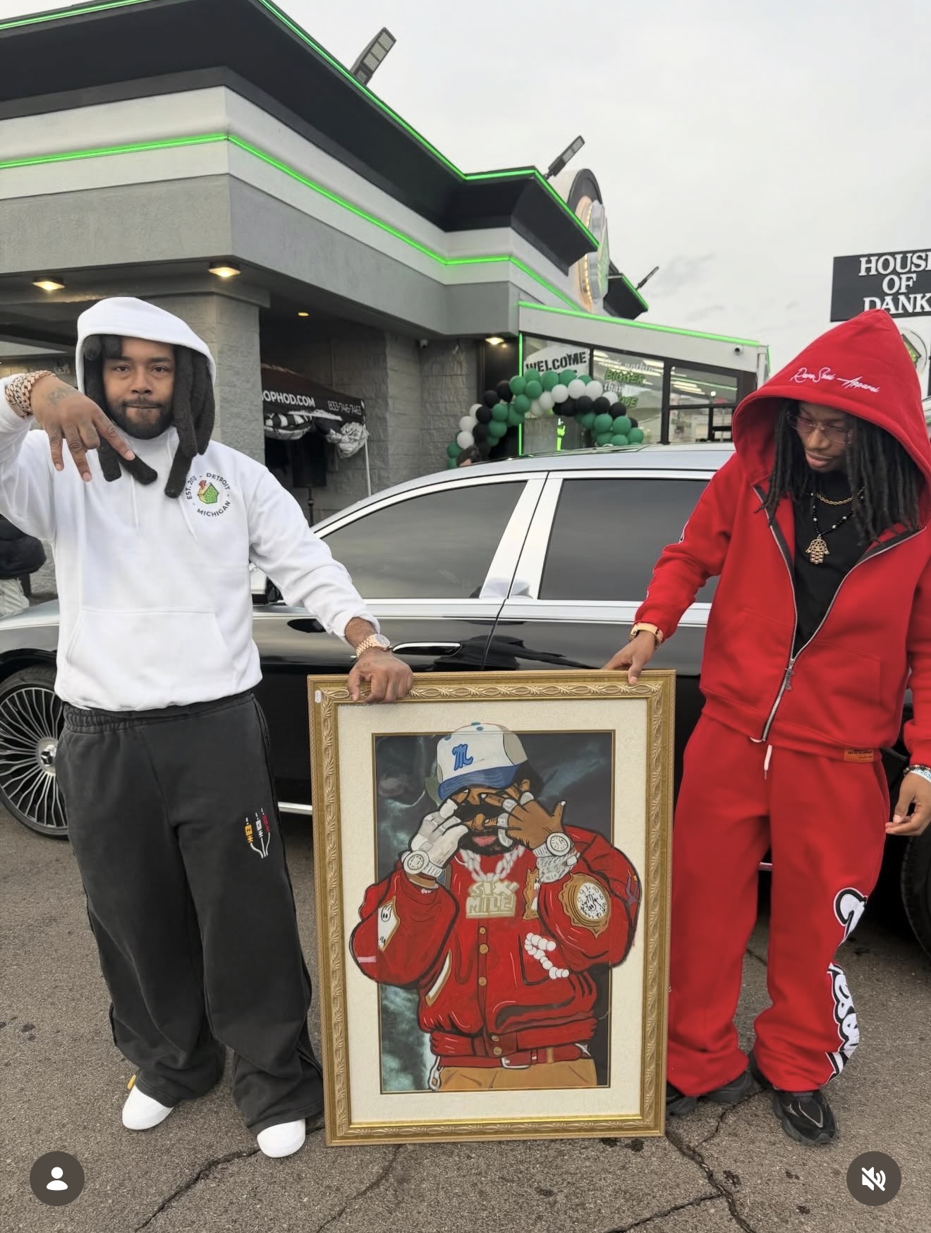 Two people stand outdoors holding a framed artwork of a cartoon character wearing a red jacket and blue hat. One person wears a white hoodie, the other a red hoodie. A car and building are in the background.