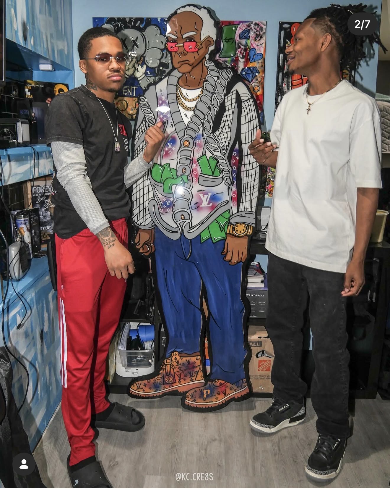 Two young men stand next to a colorful cartoon cutout of an older woman in a store, holding drinks and smiling.
