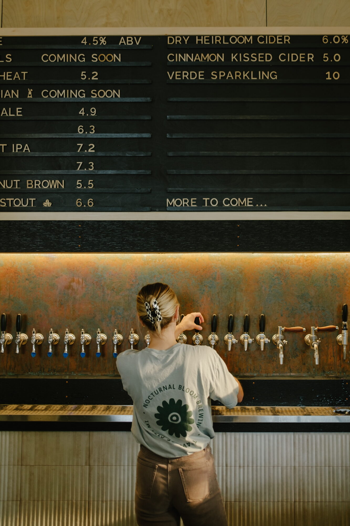 Person with a bun hairstyle and a white shirt with a green gear logo, pouring a drink at a bar with taps.