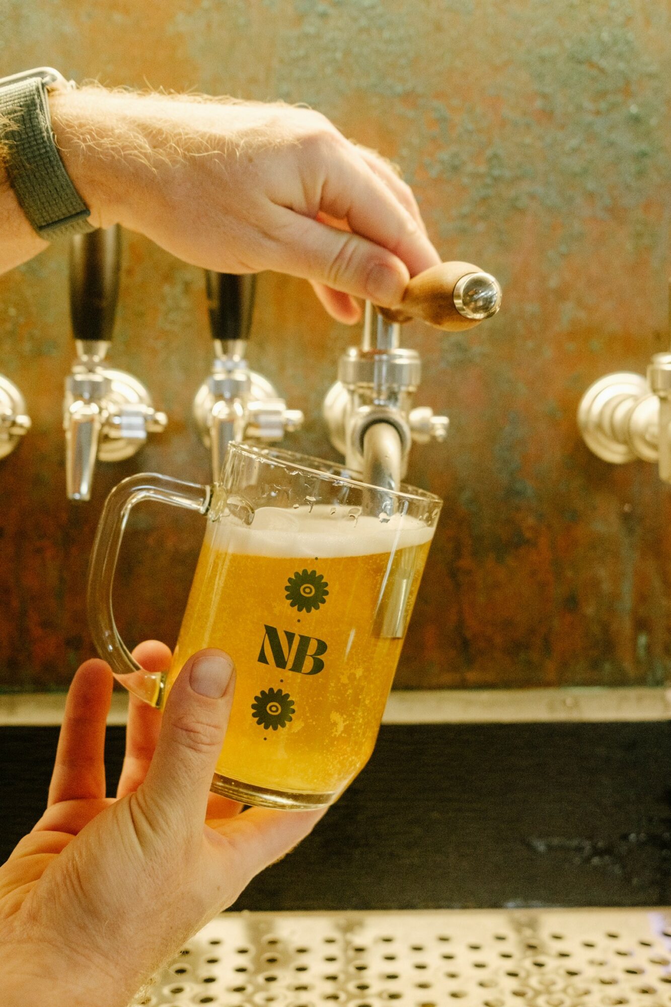 Person filling a glass with beer from a tap, with a background of textured wall and multiple taps, close-up view.