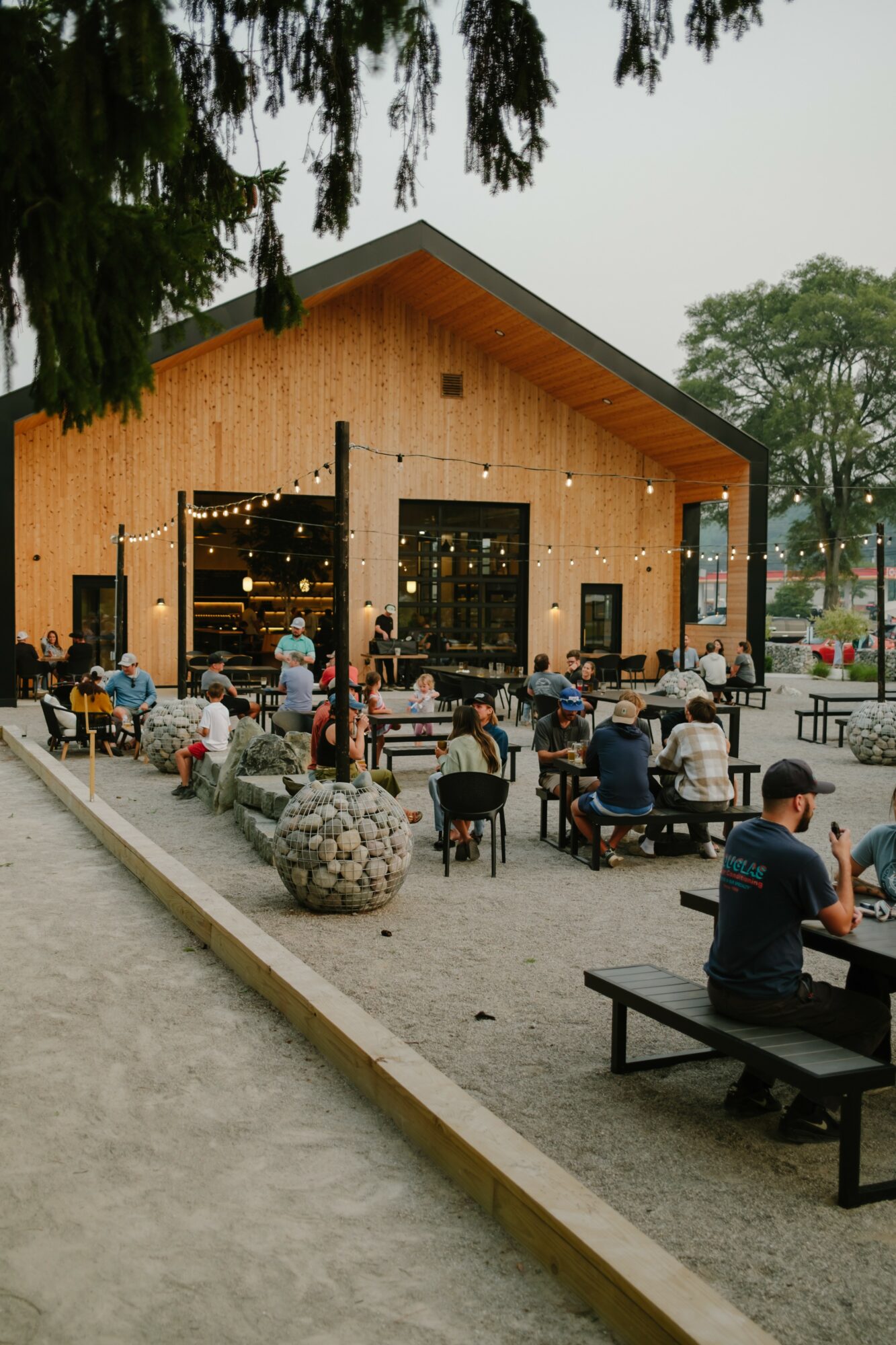 People gather outside a wooden building with string lights, sitting on benches and tables, under trees, during daytime.