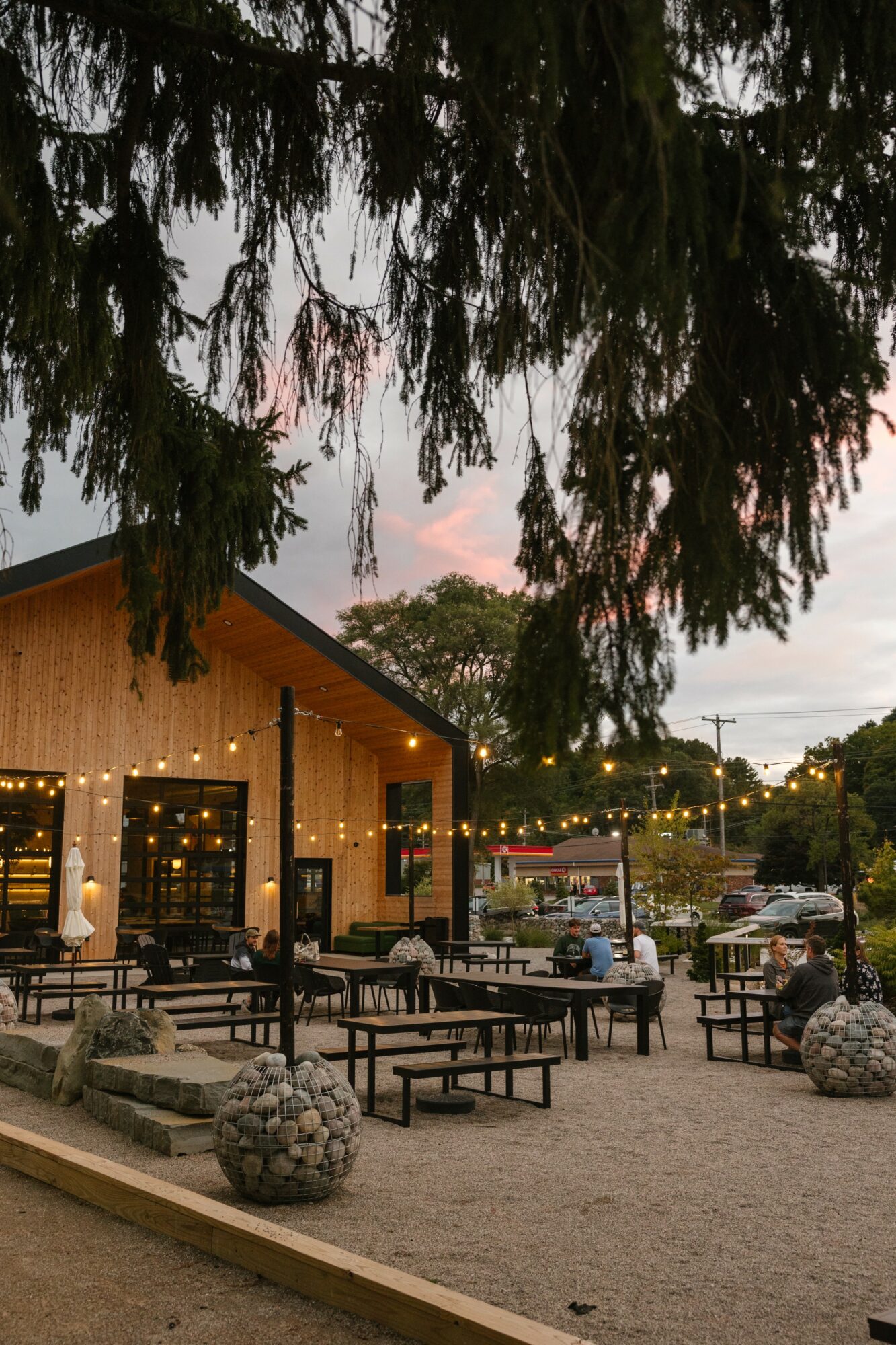 Outdoor dining area with tables and benches in front of a building, under a large tree, during sunset.