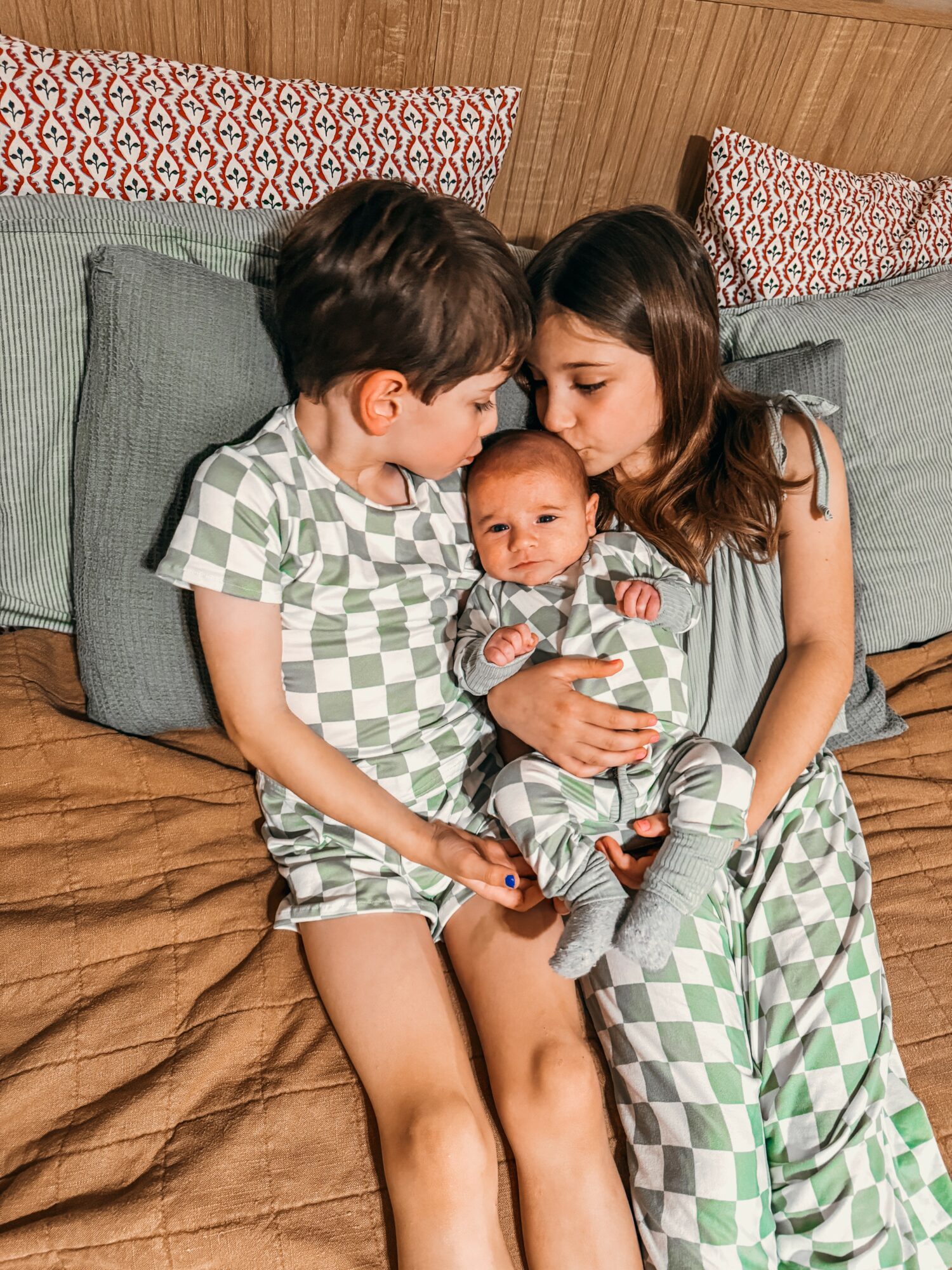 Two children and a baby sitting on a bed, with the children holding the baby, in a cozy room with pillows.