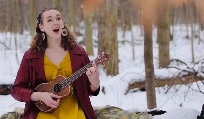 Young woman with curly hair playing a ukulele outdoors in a snowy forest, smiling and singing, wearing a yellow dress and maroon jacket.