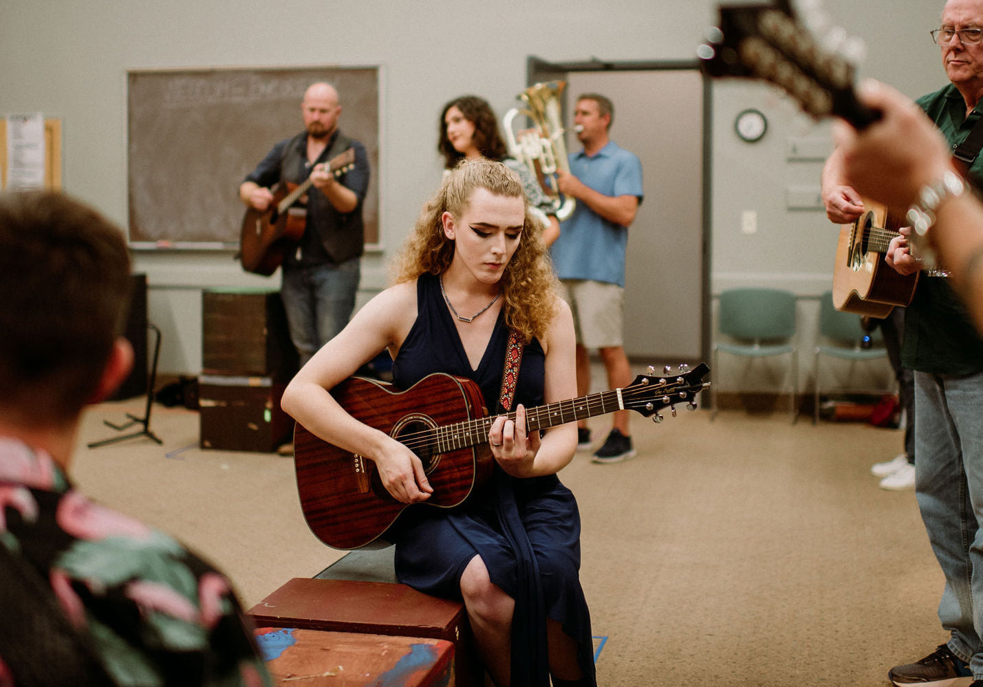 A woman plays guitar in a room with a group of people, some playing instruments, others watching.