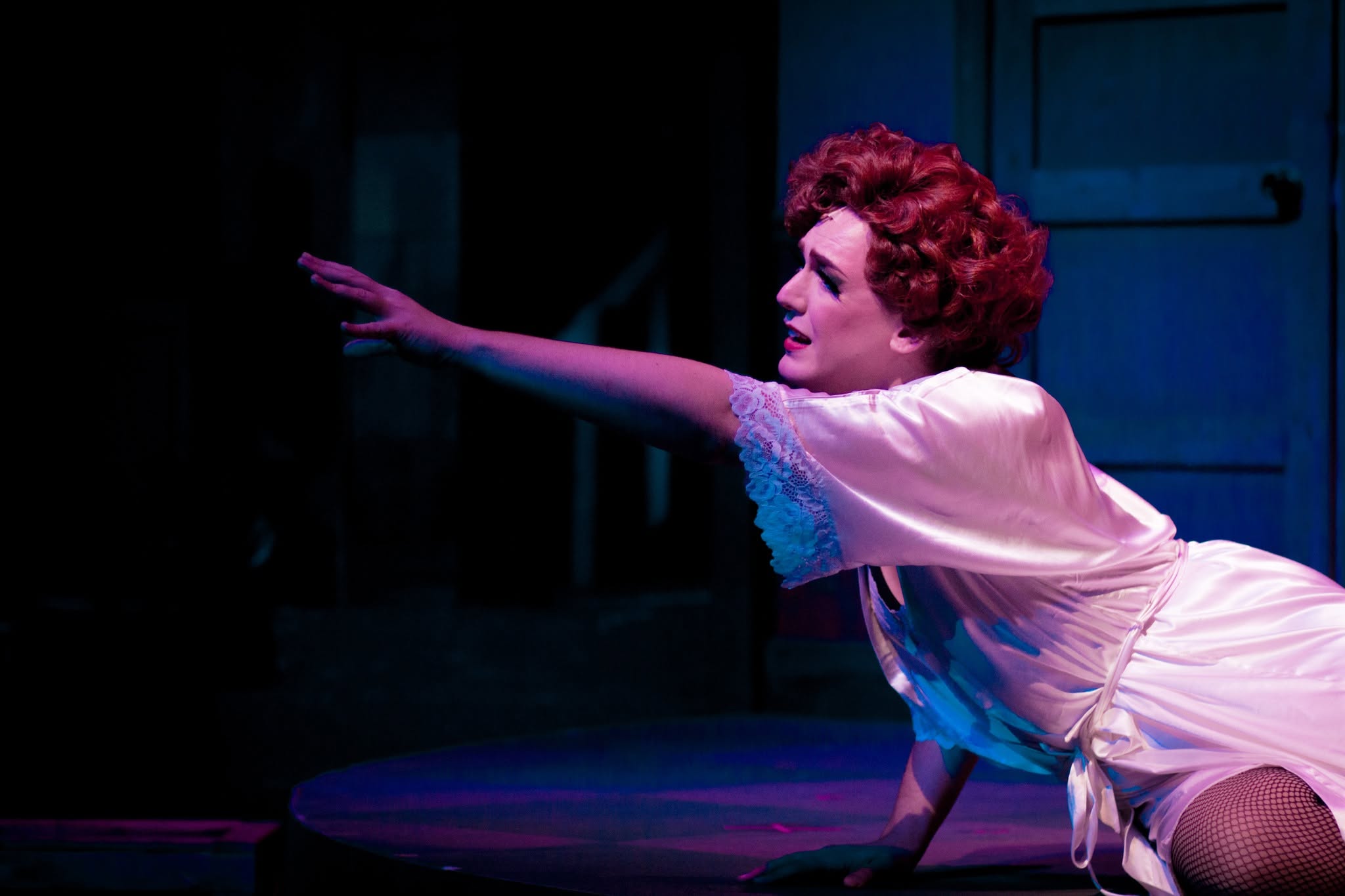 Woman with curly hair reaching forward on stage, wearing a light-colored dress, in a dark setting with blue lighting.