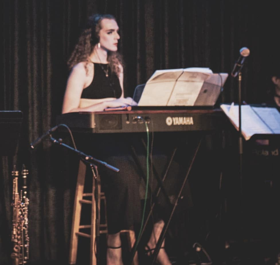 Woman playing keyboard on stage with music sheet, microphone, and dark curtain background.