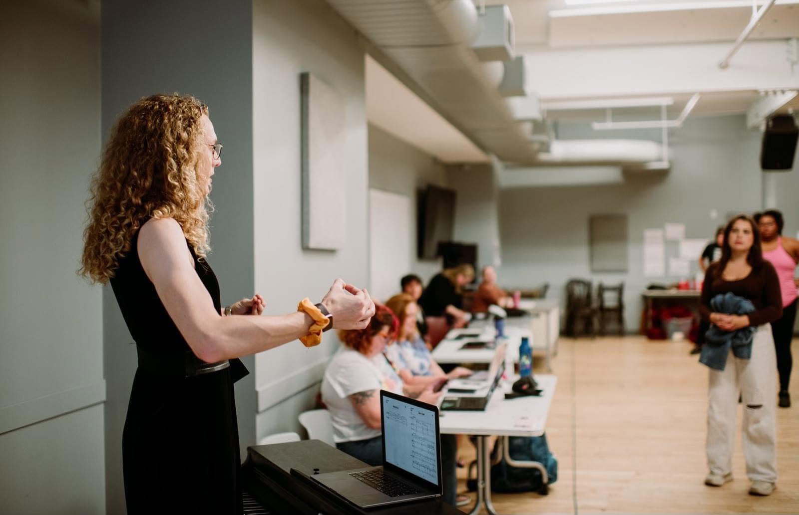 A woman with curly hair holding a microphone in a room with people sitting at tables using laptops.