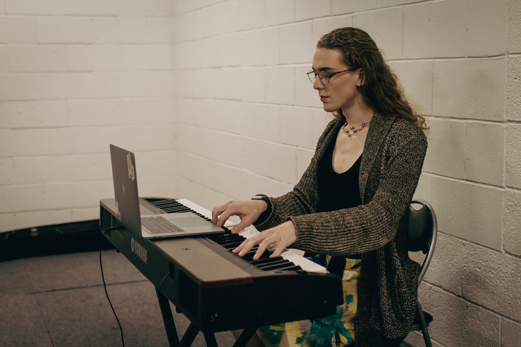 Woman playing keyboard with laptop on a stand, against a white brick wall, wearing glasses and a cardigan.