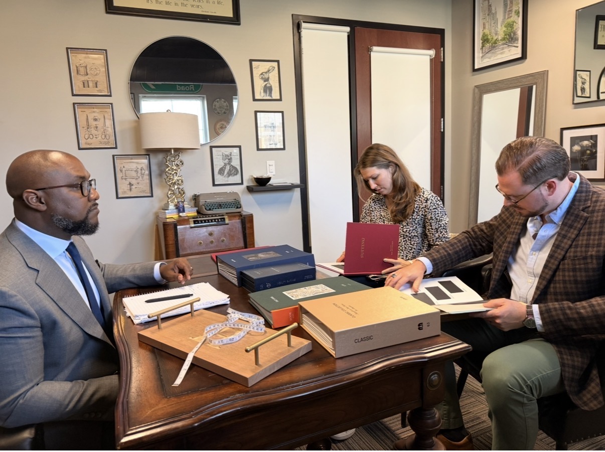 Three people sit at a table with files and documents in a room with framed pictures and a mirror on the wall.