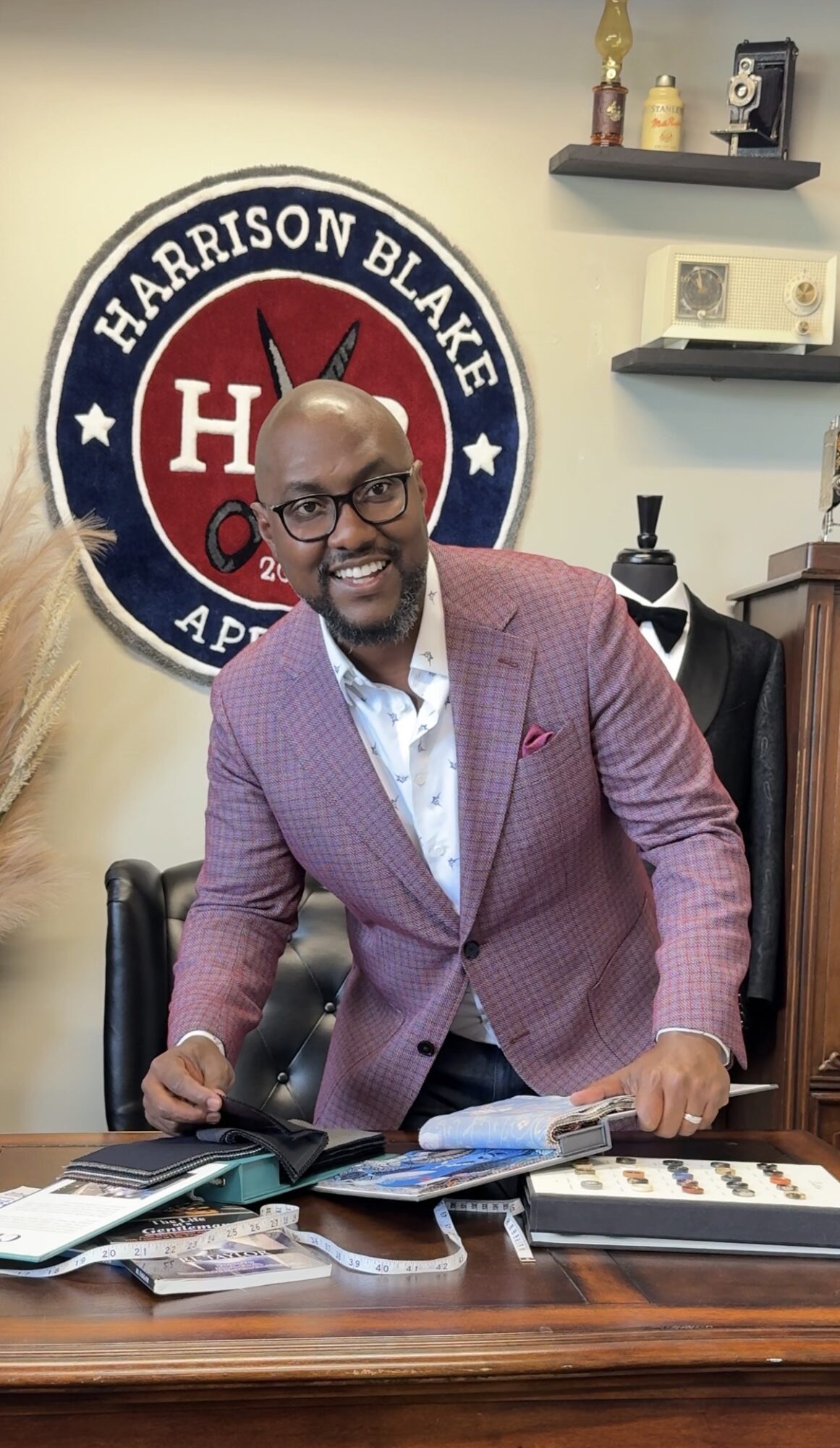 Man in a pink blazer and glasses leaning on a desk with papers, in front of a Harrison Blake sign.
