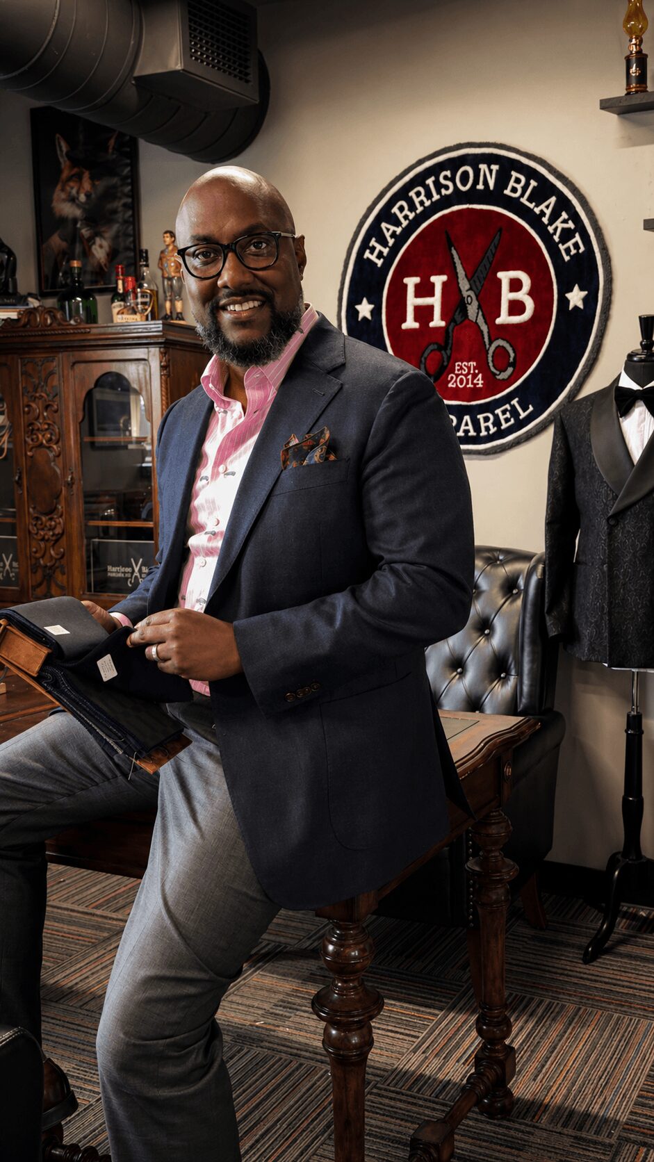 Man with glasses in a suit holding a book, standing in a room with vintage furniture and Harrison Blake logo.