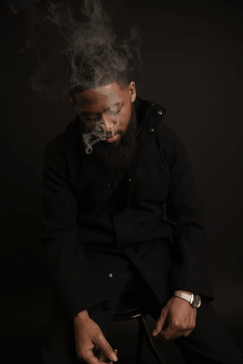 Man with a beard and glasses smokes, with smoke rising from his mouth, wearing a black jacket against a dark background.