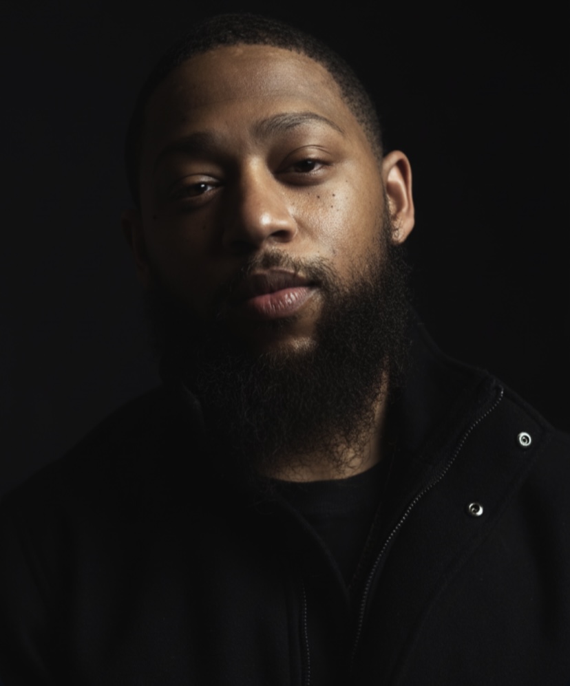 Portrait of a man with a beard and short hair, wearing a dark jacket, against a dark background.