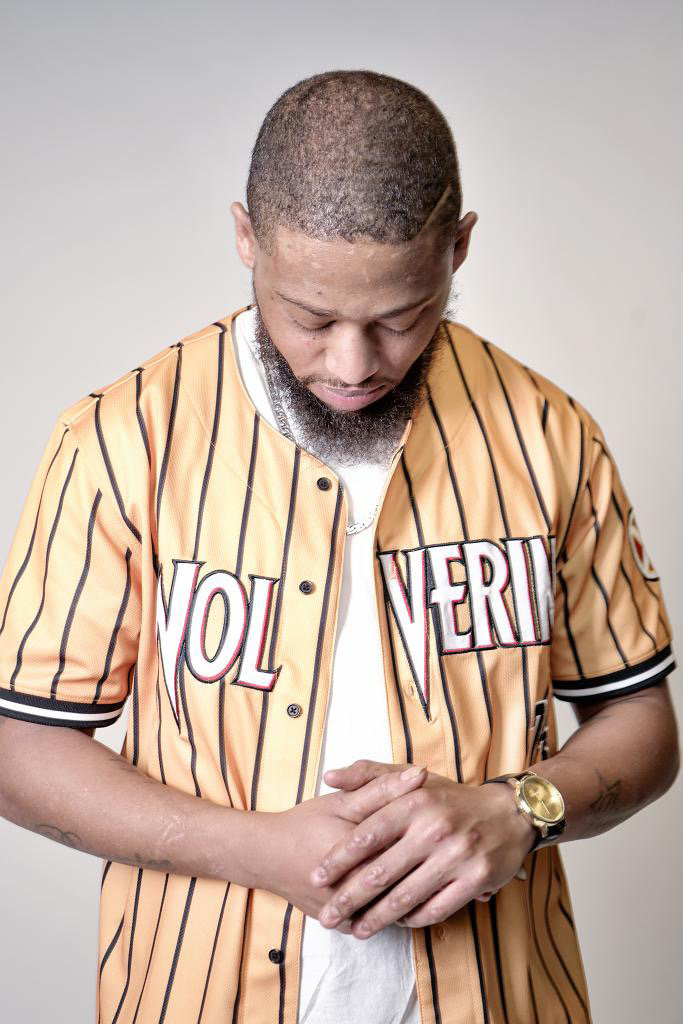 Man with short hair and beard wearing a striped shirt, looking down with hands clasped, against a plain background.