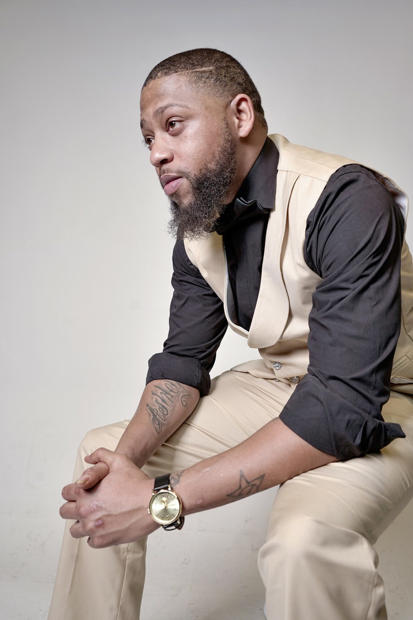 Man with a beard and short hair, wearing a black shirt and beige vest, sitting with hands clasped, looking to the side.