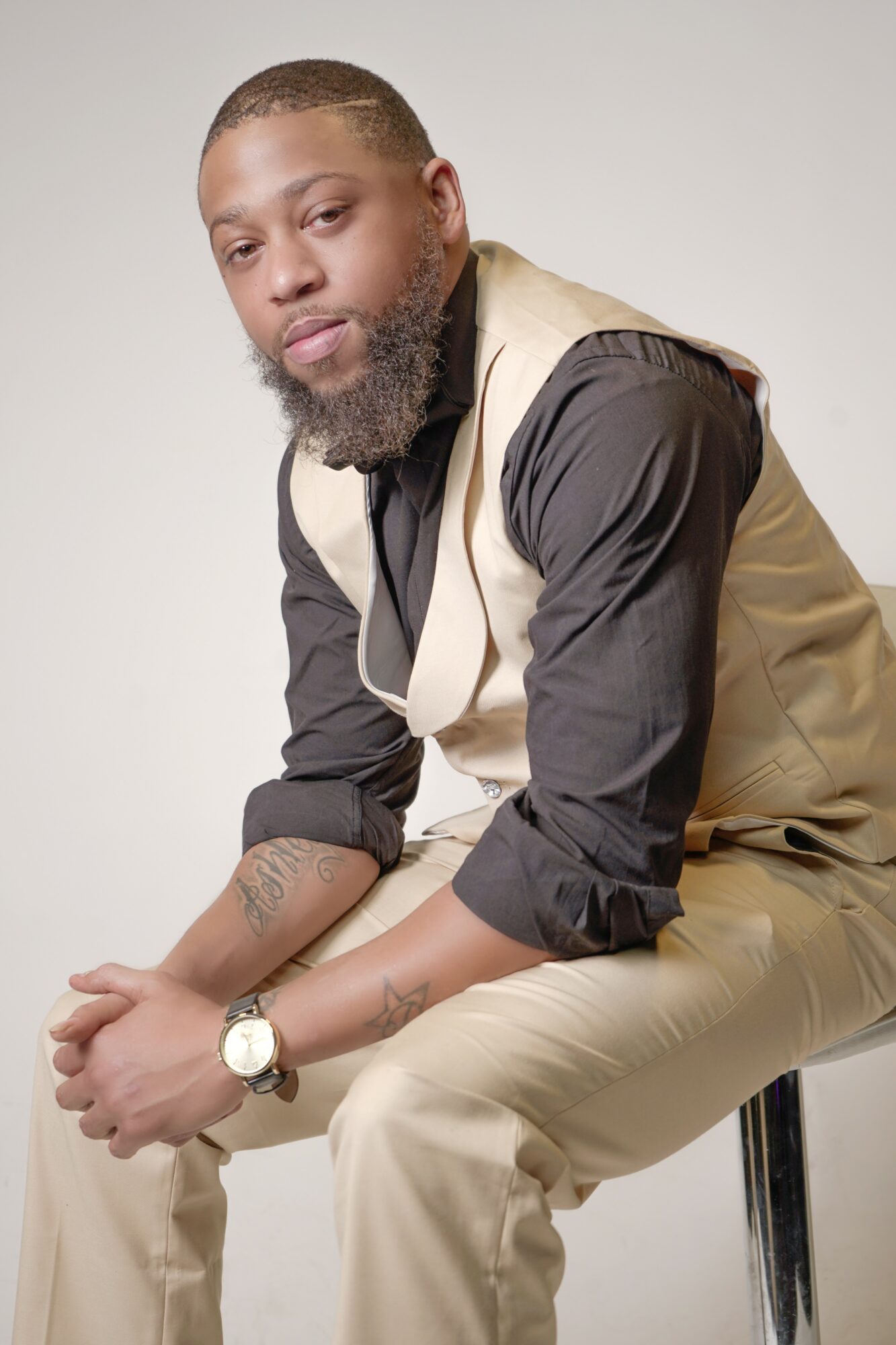 Man with a beard and tattoos, wearing a beige vest over a black shirt, sitting on a stool against a plain background.