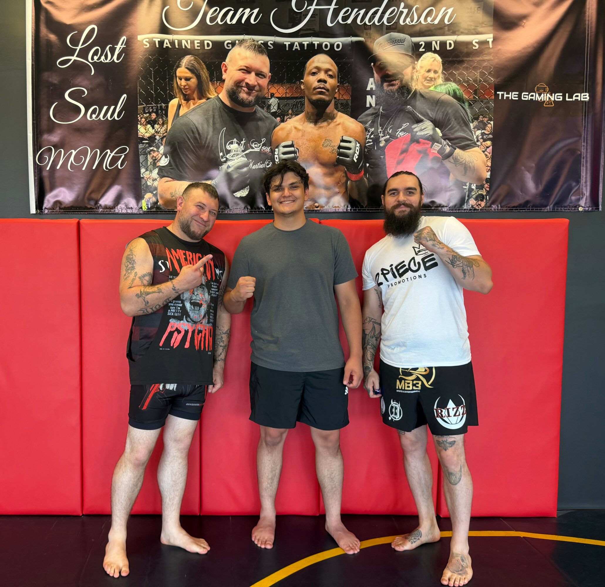 Three men stand in front of a red wall, with a large poster of fighters behind them, in a gym setting.
