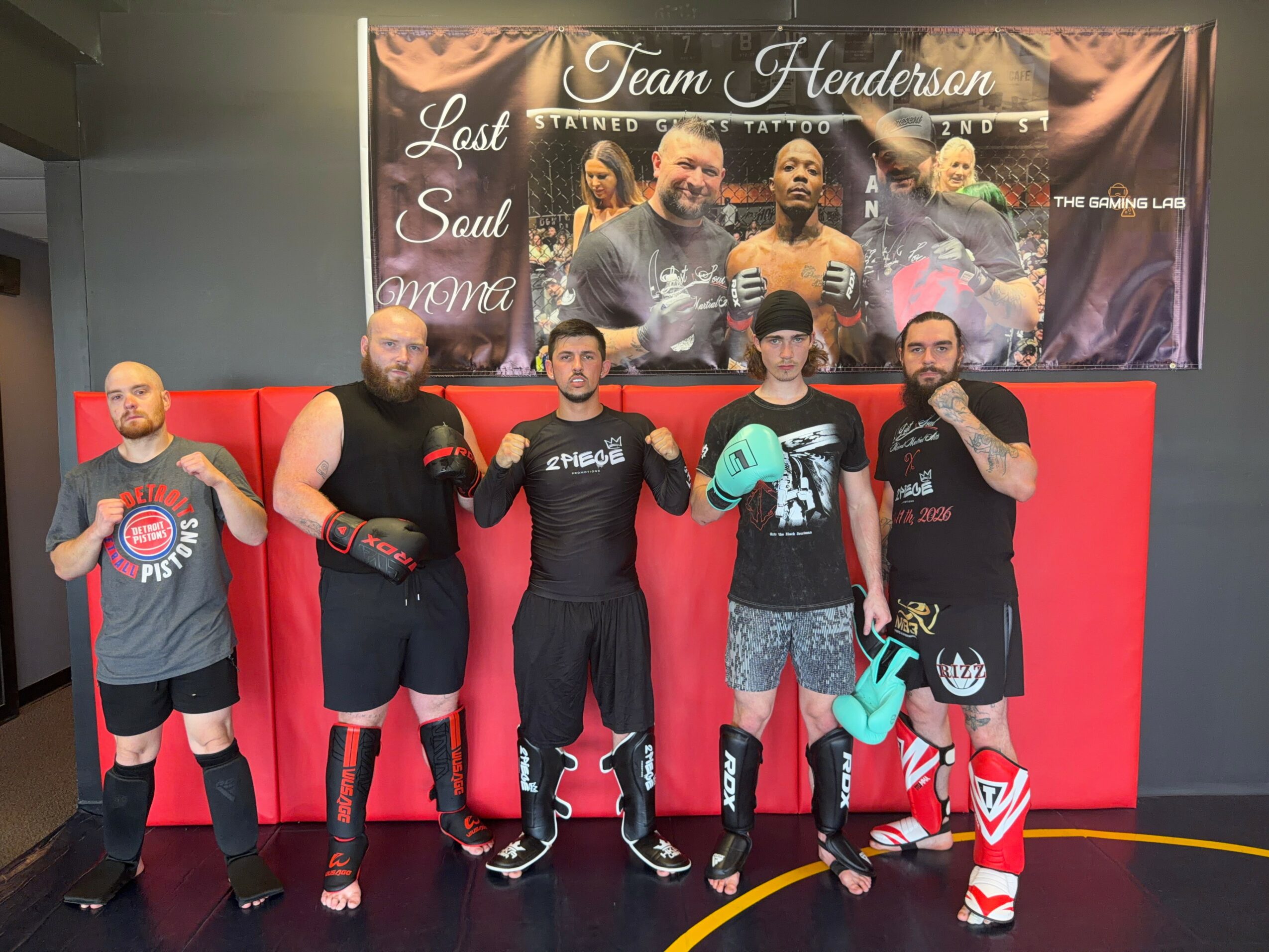 Five men in athletic clothing and boxing gloves stand in front of a red padded wall, holding boxing gloves, with a banner above them.