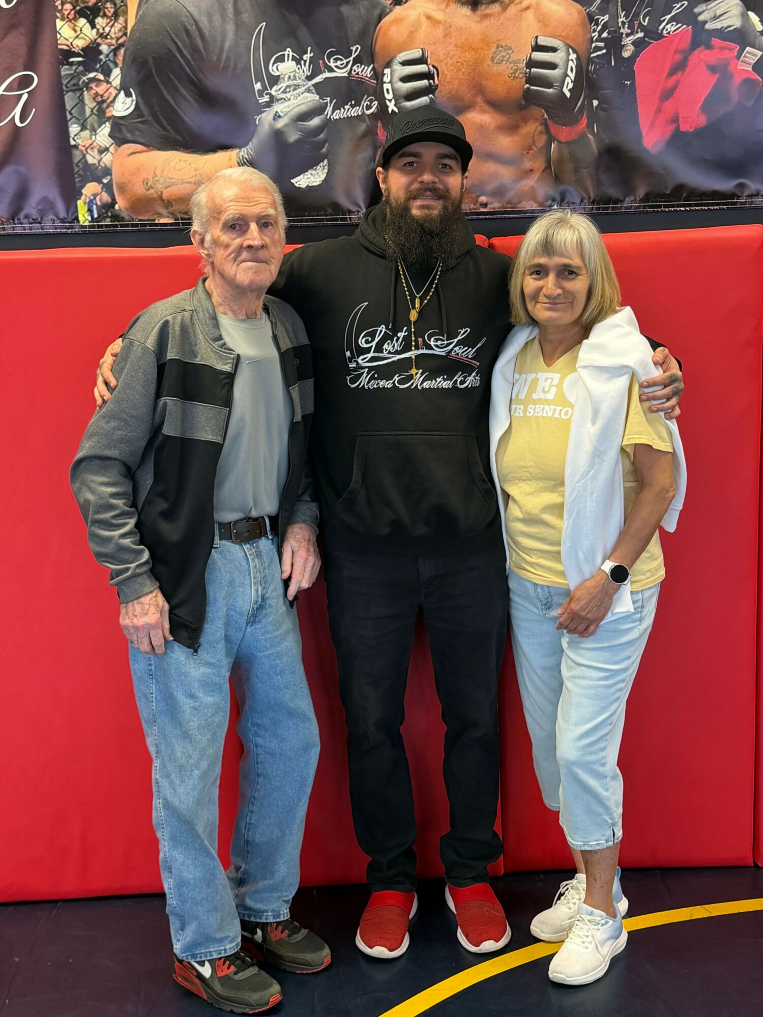Three people standing together in front of a red background, smiling, with a dog and promotional posters behind them.