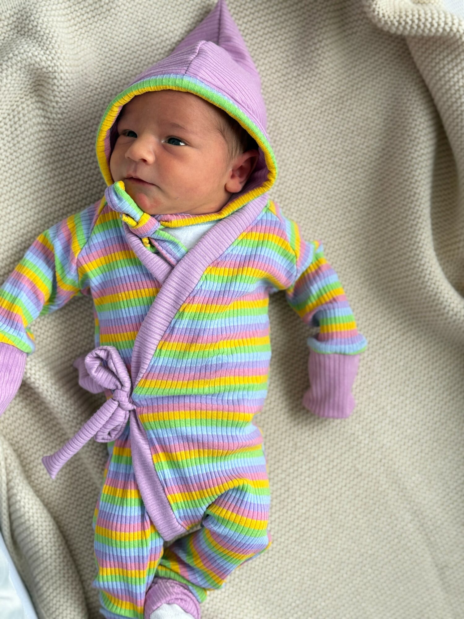 Baby in colorful striped outfit with purple hat lying on a cream blanket.