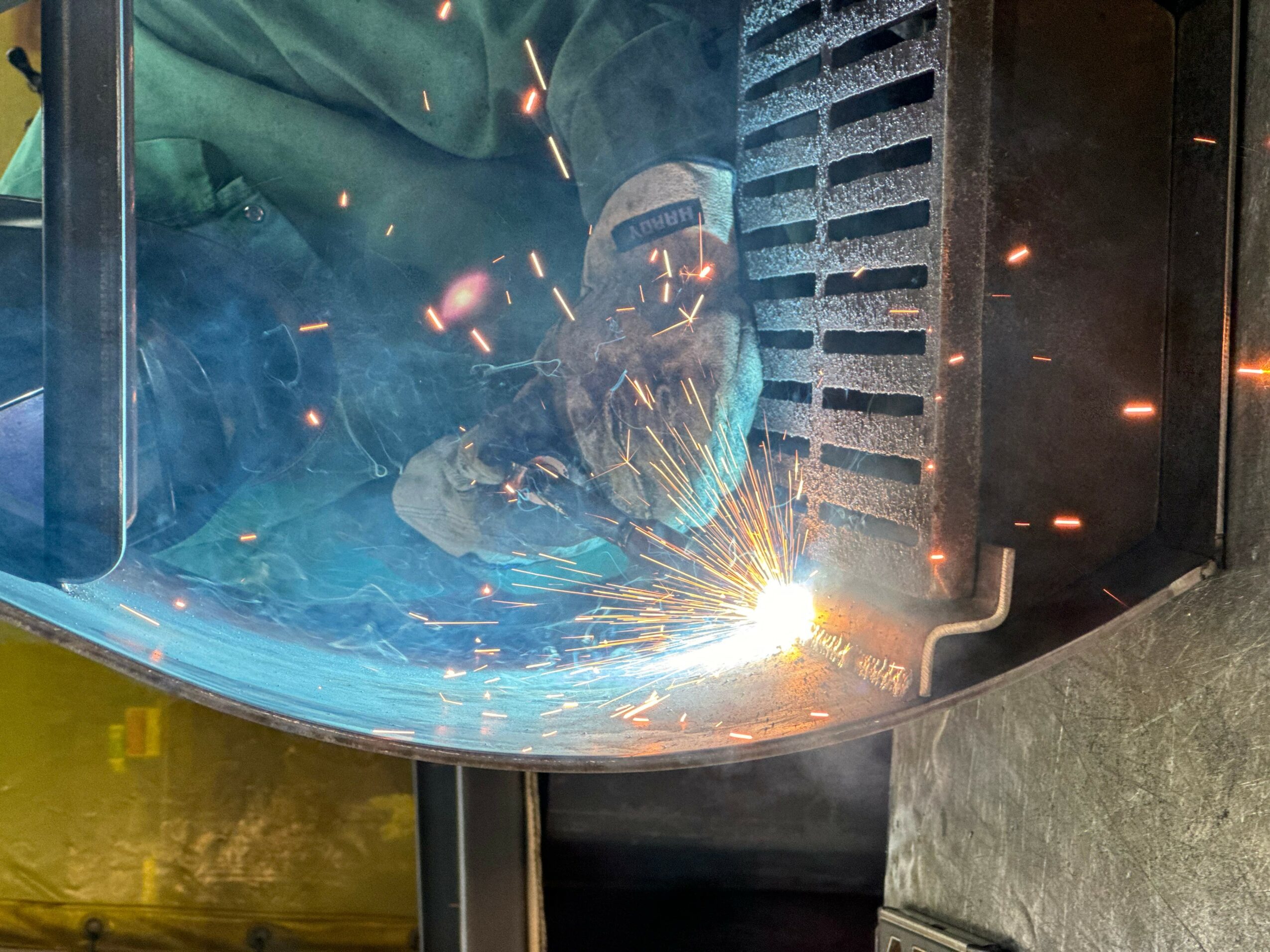 Welder wearing protective gear welding metal with sparks flying, inside a workshop or industrial setting.