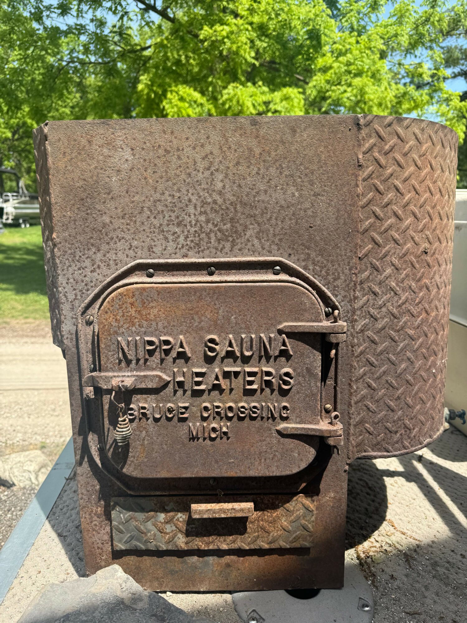 A metal mailbox with a rounded top, mounted on a rusted metal surface, with trees and a street in the background.