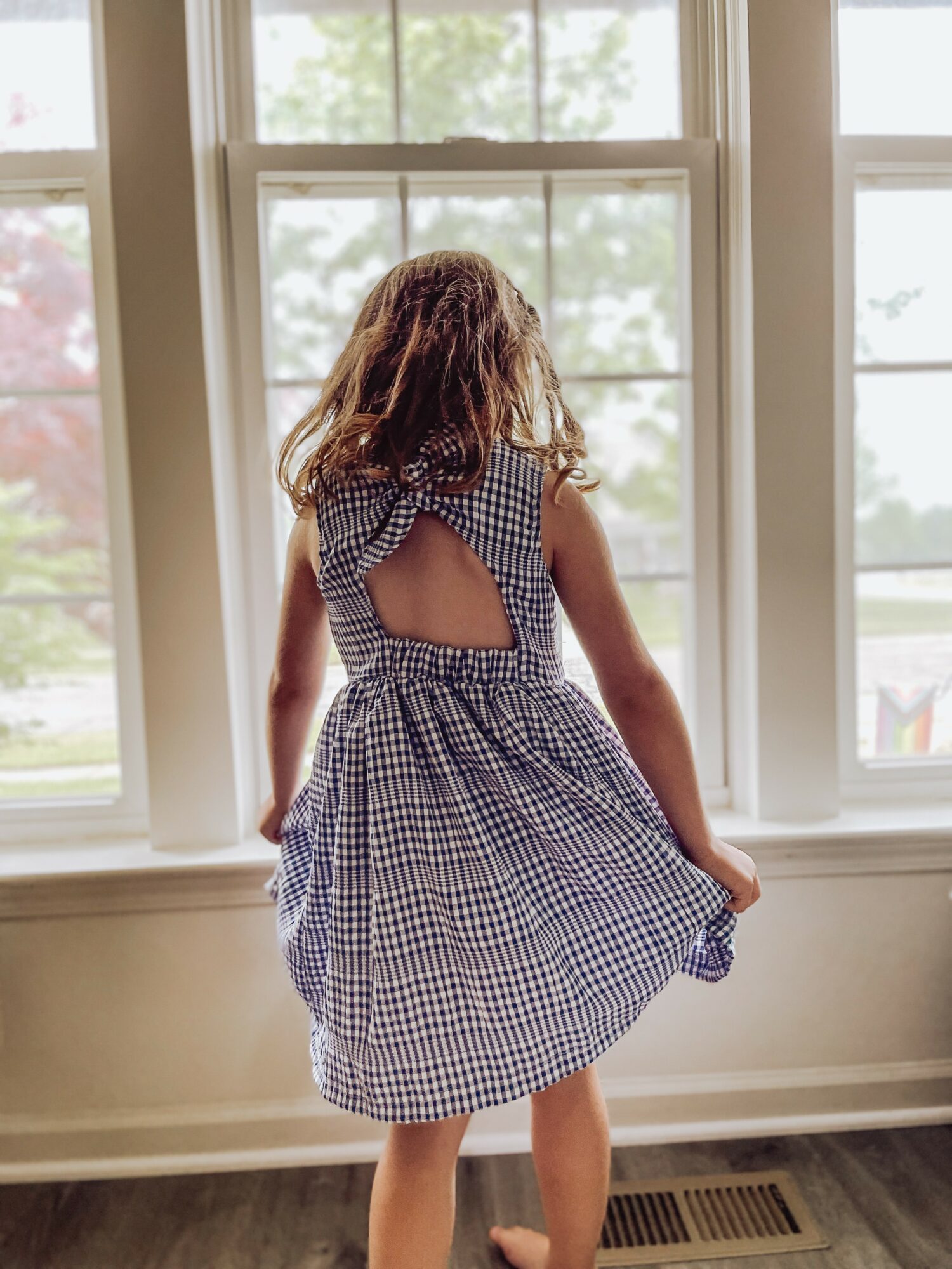 Child with curly hair in a checkered dress standing by a large window, looking outside, with natural light illuminating the scene.