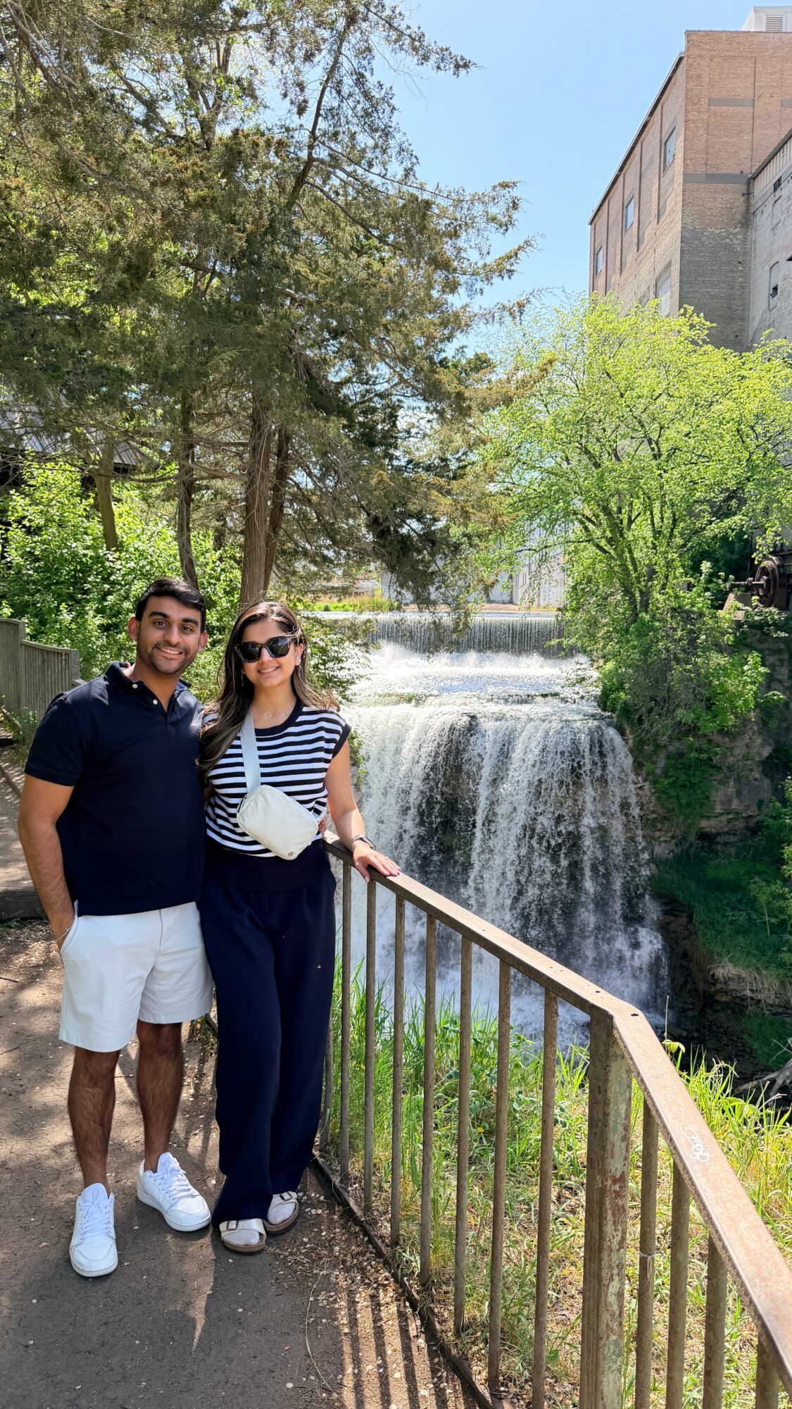 Two people stand by a railing near a waterfall with trees and a building in the background.