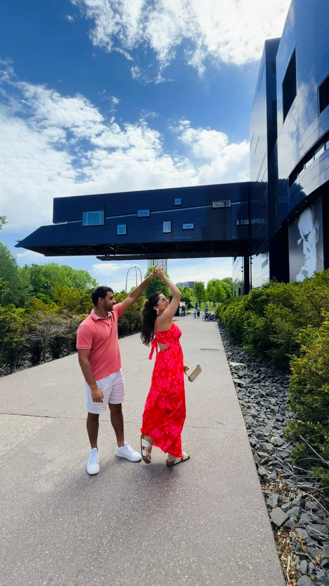 Two people standing on a sidewalk near a modern building with a blue sky and clouds above.