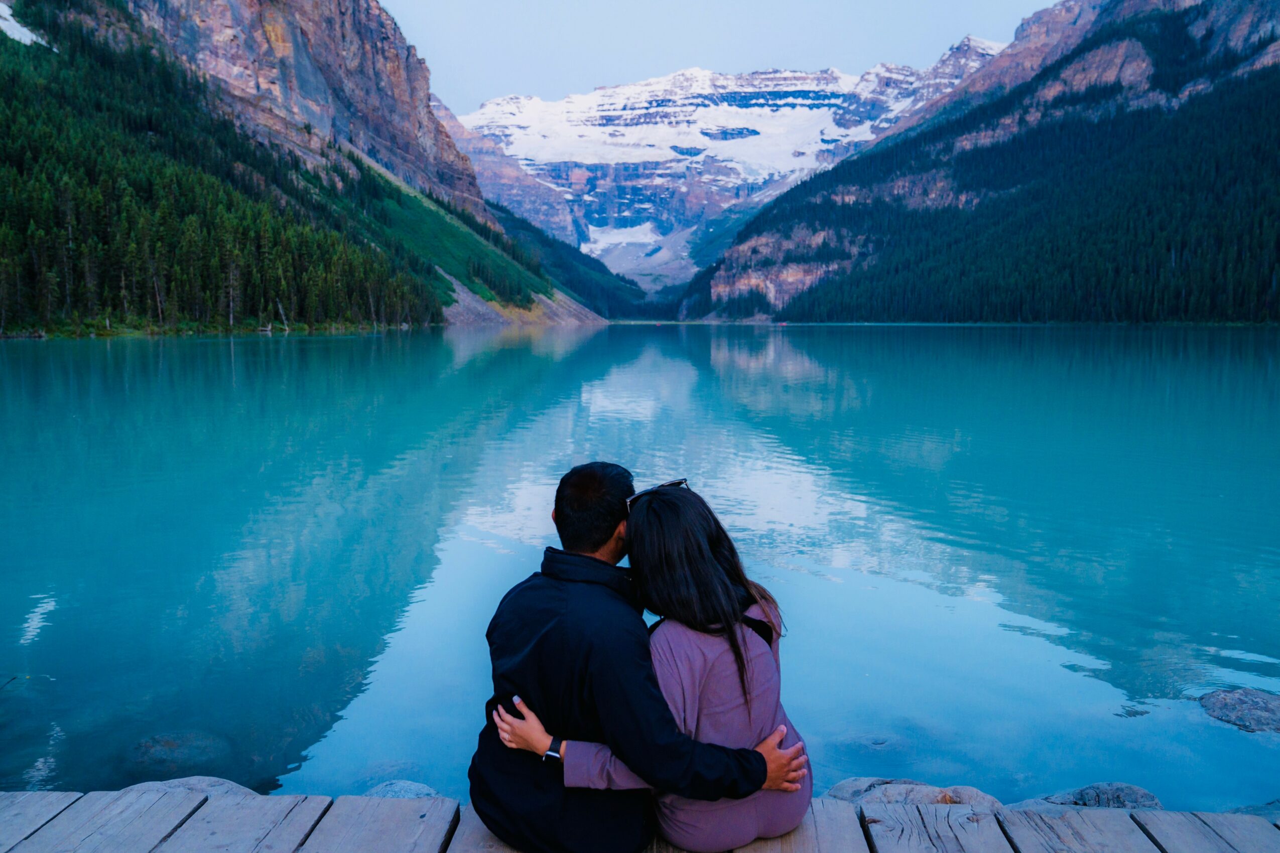 Two people sit on rocks by a turquoise lake surrounded by mountains and snow-capped peaks, with trees along the shoreline.