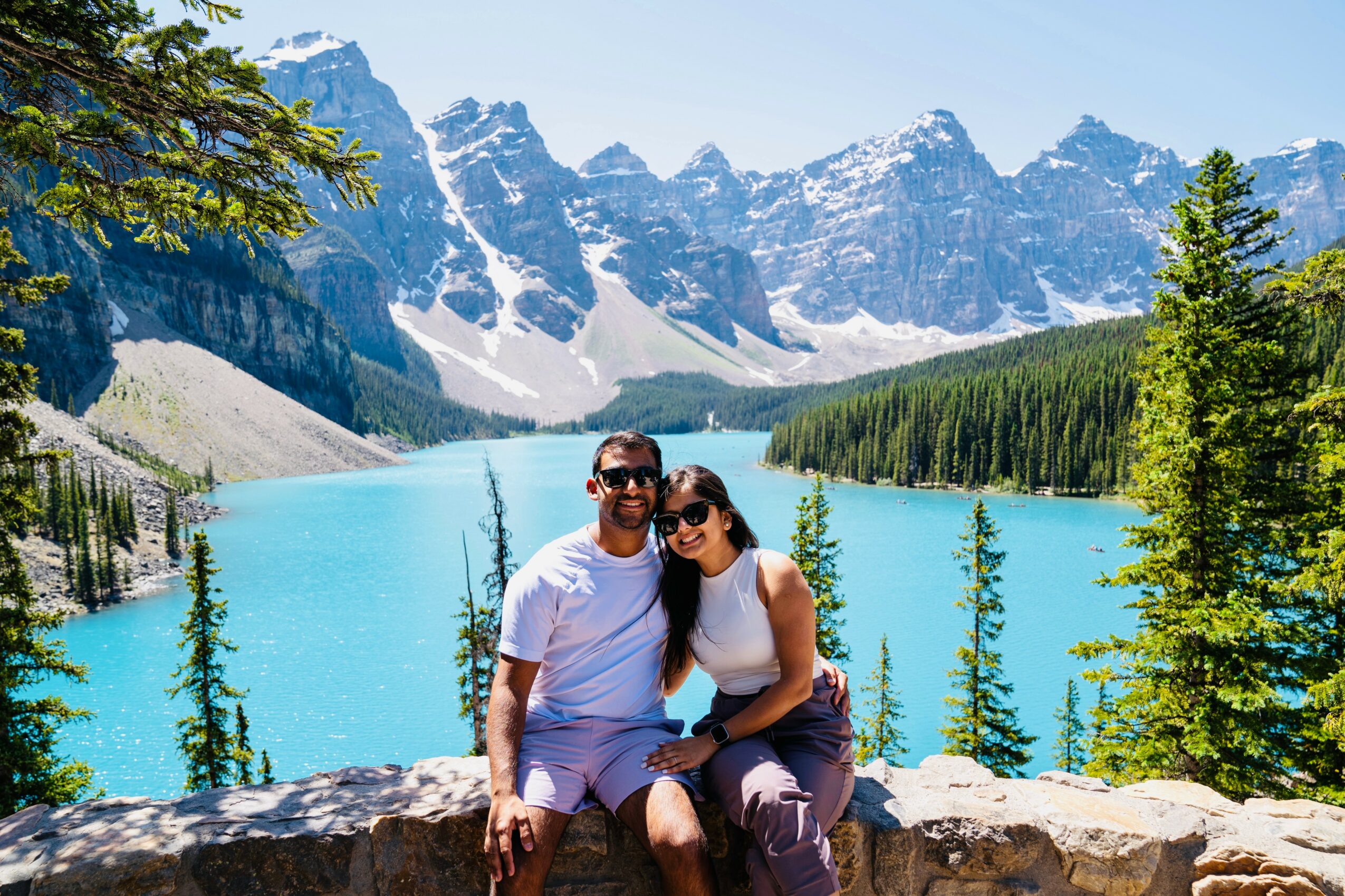 A couple sitting on a rock in front of a turquoise lake with mountains and trees in the background.